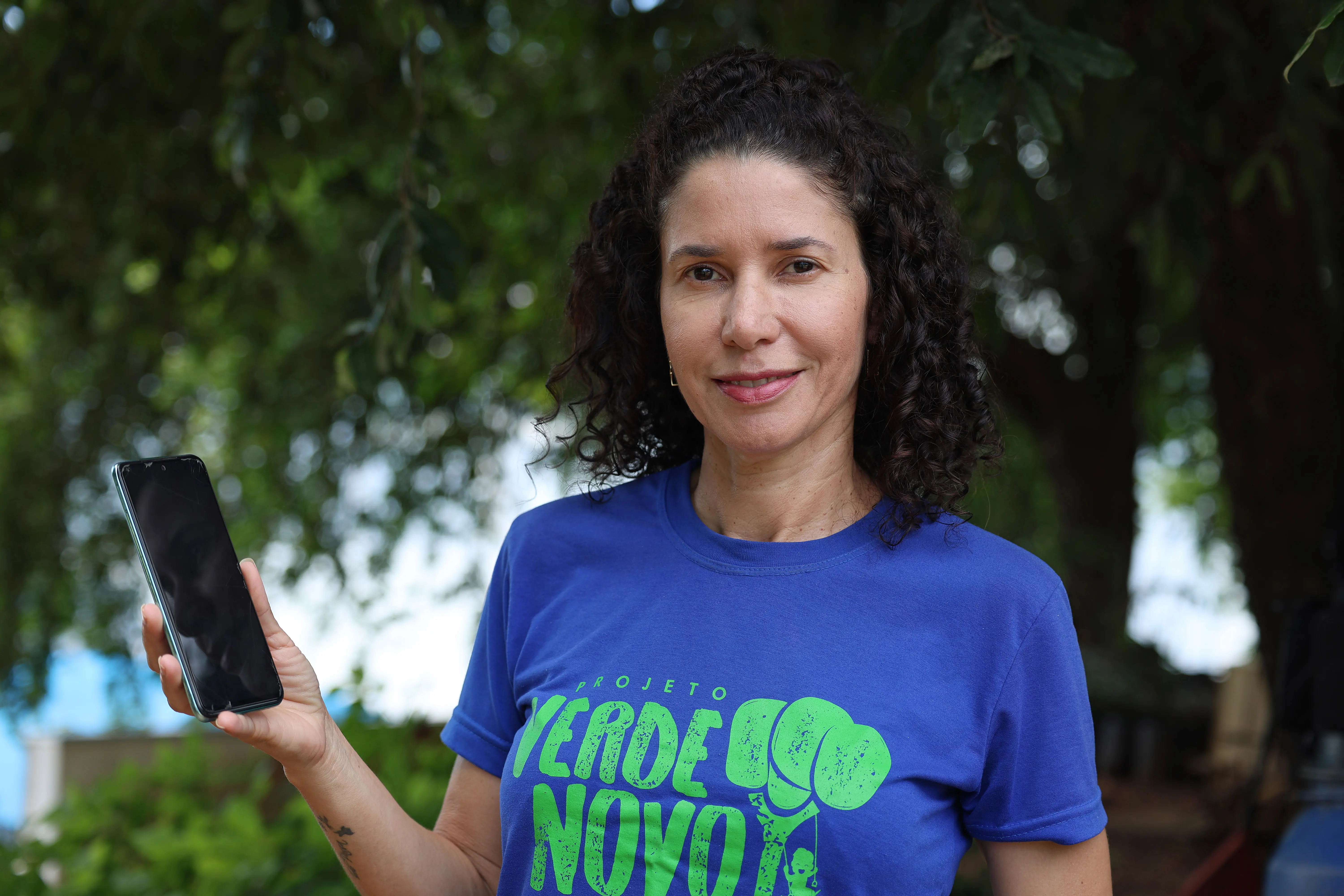 Mulher de cabelos cacheados veste camiseta azul com a logo "Projeto Verde Novo" e segura um smartphone. Ao fundo, há vegetação densa e sombras de árvores.