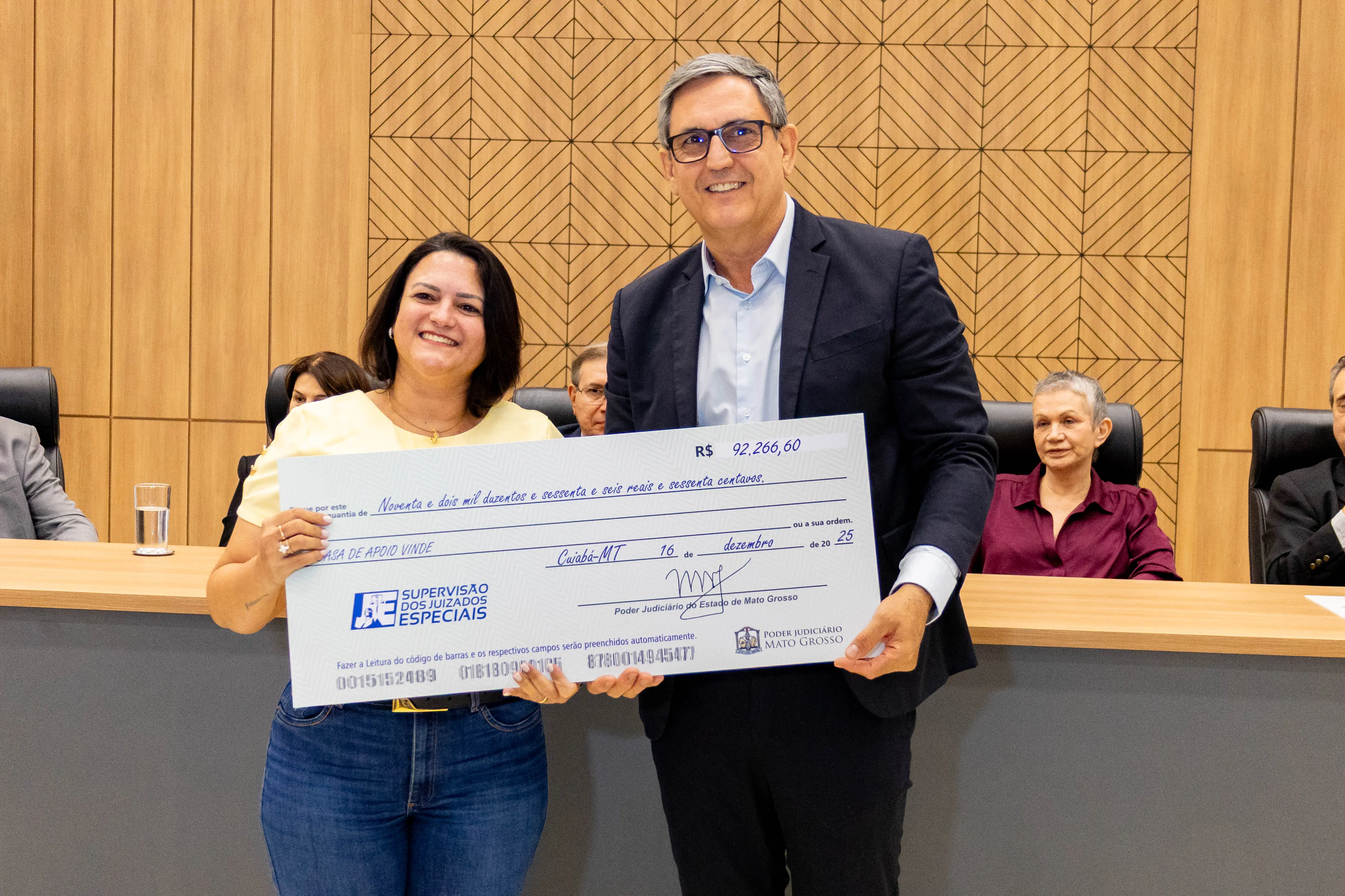 Mulher e homem posam sorrindo, segurando cheque simbólico durante solenidade no plenário. Ao fundo, cadeiras do colegiado e ambiente institucional iluminado.