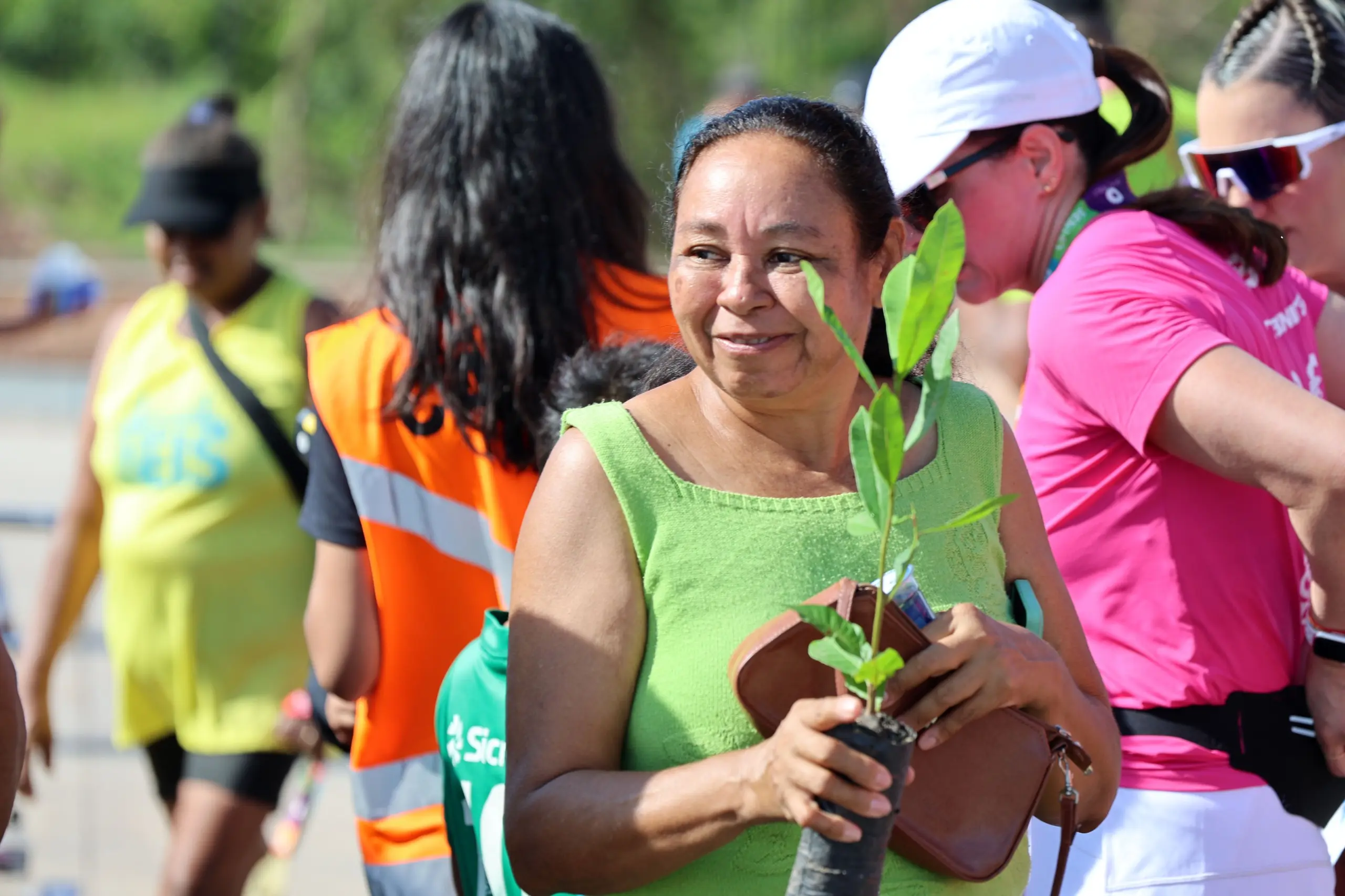 Mulher de vestido verde segura uma muda de árvore junto ao corpo e observa o movimento ao redor. Ao fundo, participantes com camisetas coloridas e coletes circulam pela área externa da corrida, em ambiente ensolarado.