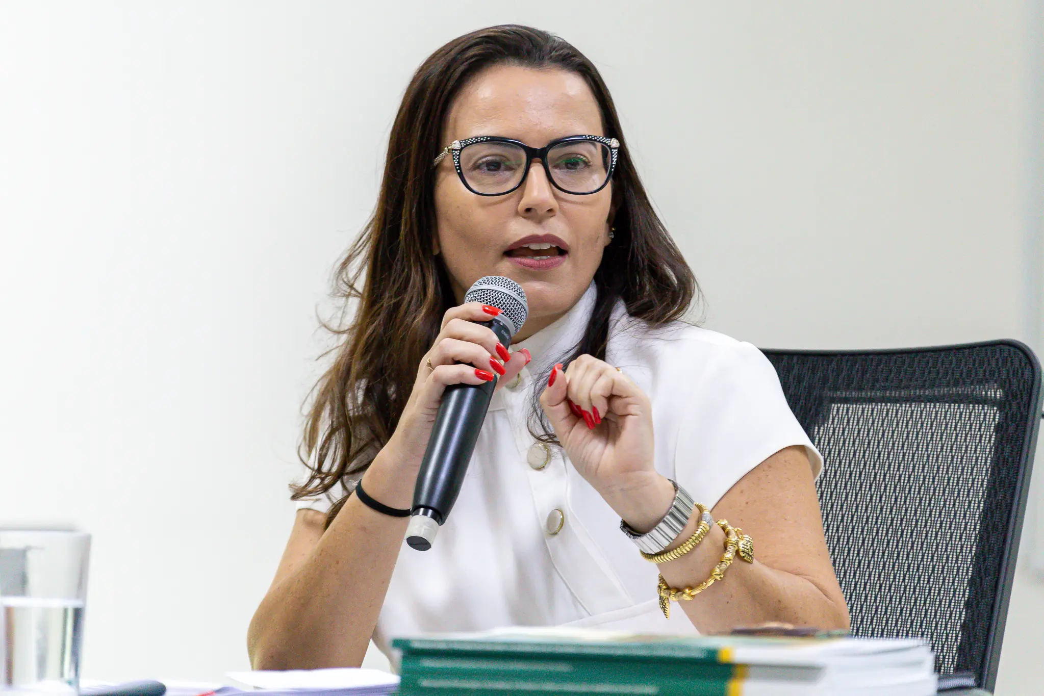 Juíza Emanuelle tem pele clara, cabelos castanhos longos e óculos, sentada em ambiente interno, fala ao microfone durante evento institucional. Usa blusa branca e acessórios dourados. À frente, há mesa com documentos; o espaço é formal e iluminado.