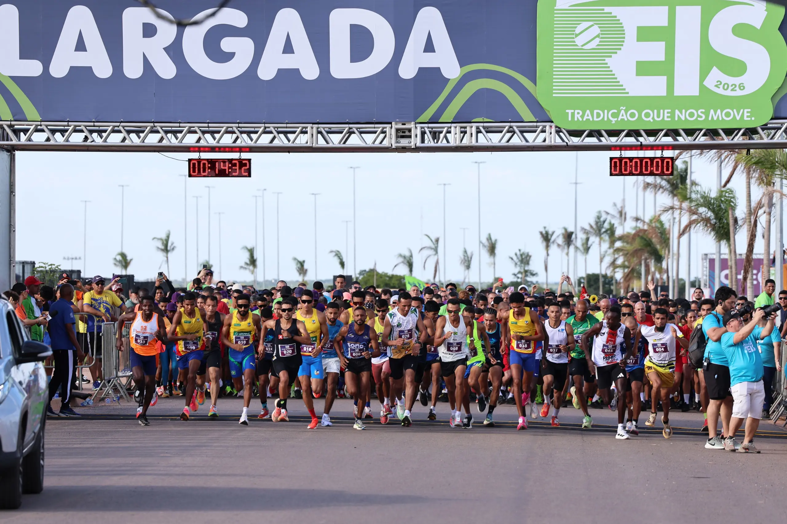 Grande grupo de corredores se concentra sob o pórtico de largada, com camisetas coloridas e números de peito. A imagem mostra o momento coletivo antes da prova, em espaço amplo e aberto.