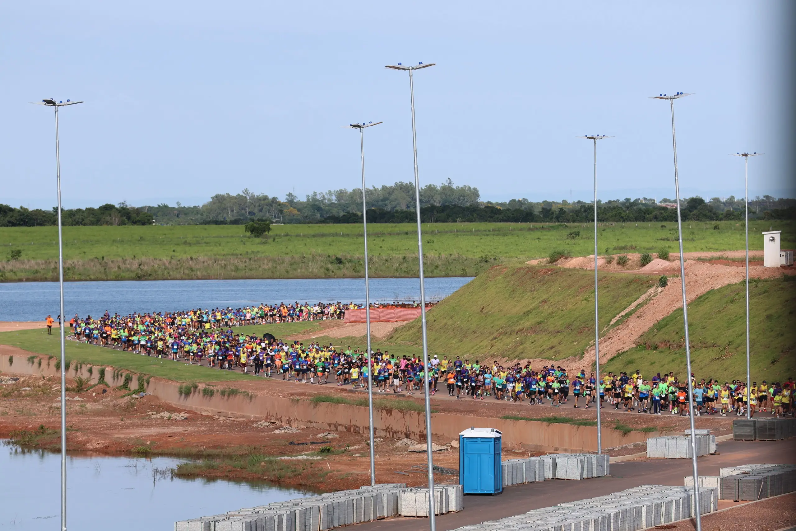 Vista aérea mostra o trajeto da corrida com corredores espalhados pela pista. O percurso passa por área aberta, com vegetação lateral e ampla visibilidade.