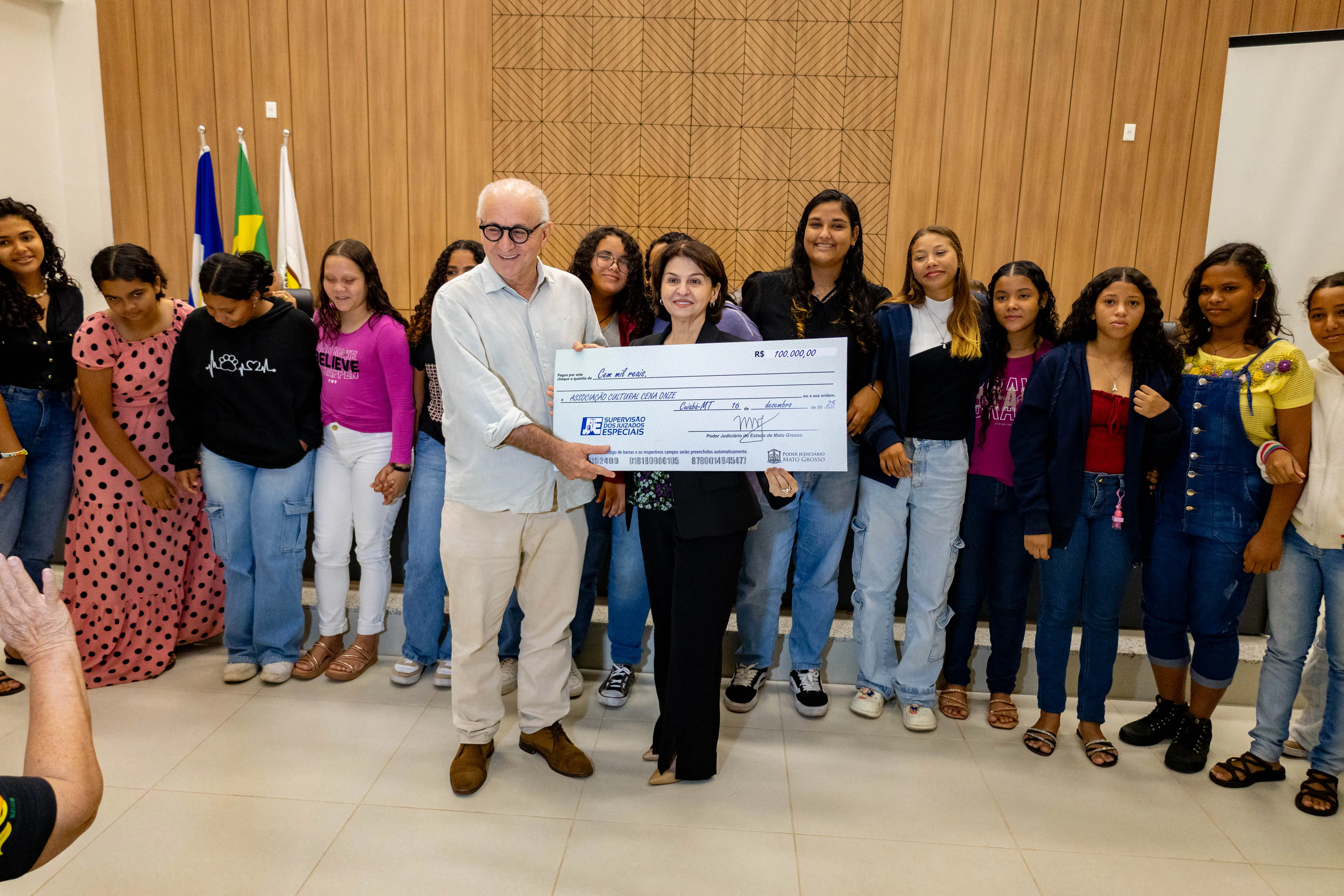 Grupo numeroso de jovens e adultos reunidos em palco, com duas pessoas ao centro segurando cheque simbólico. O ambiente é institucional, com painel de madeira ao fundo e clima de celebração coletiva.
