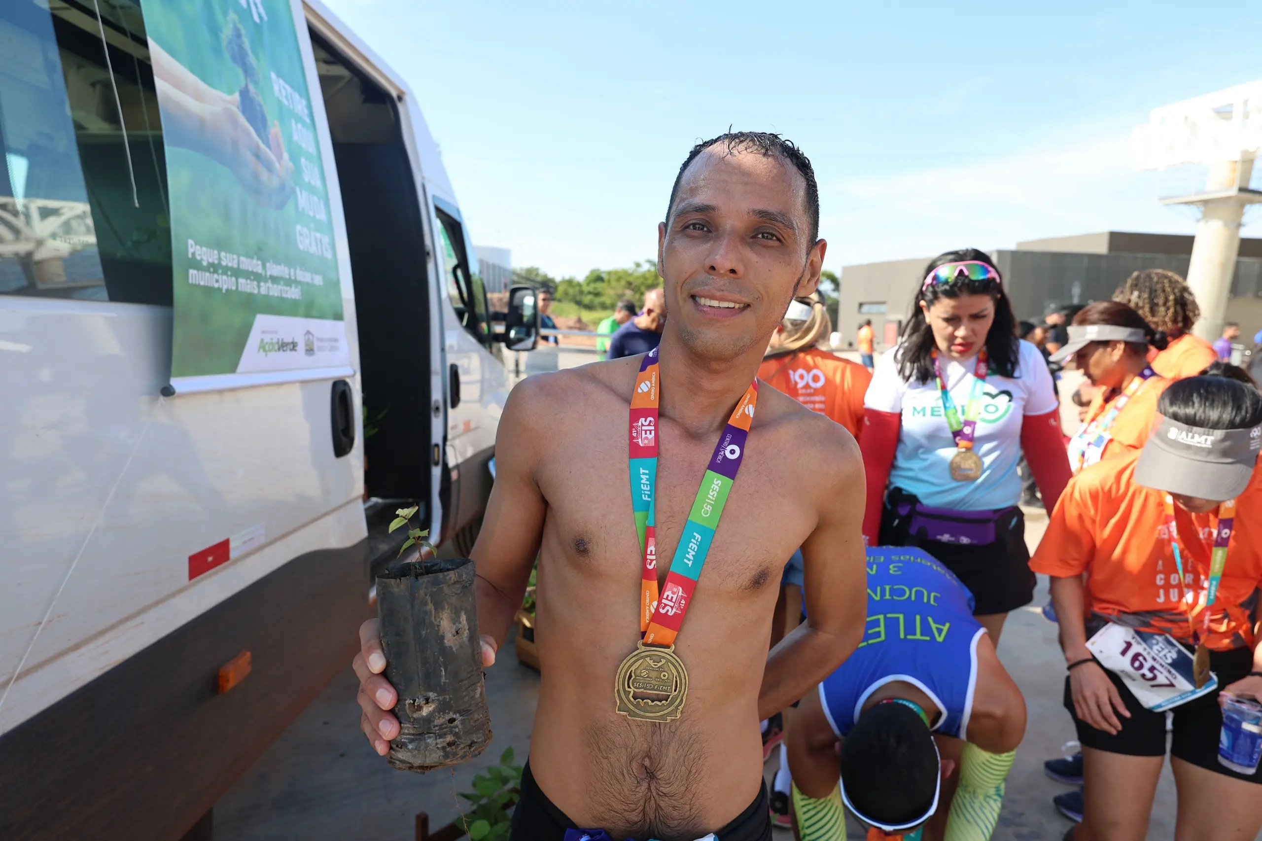 Homem sem camisa posa segurando uma muda de árvore e exibindo medalha no peito. Ele está ao lado de um veículo identificado com ação ambiental, enquanto outros corredores organizam mudas ao fundo.