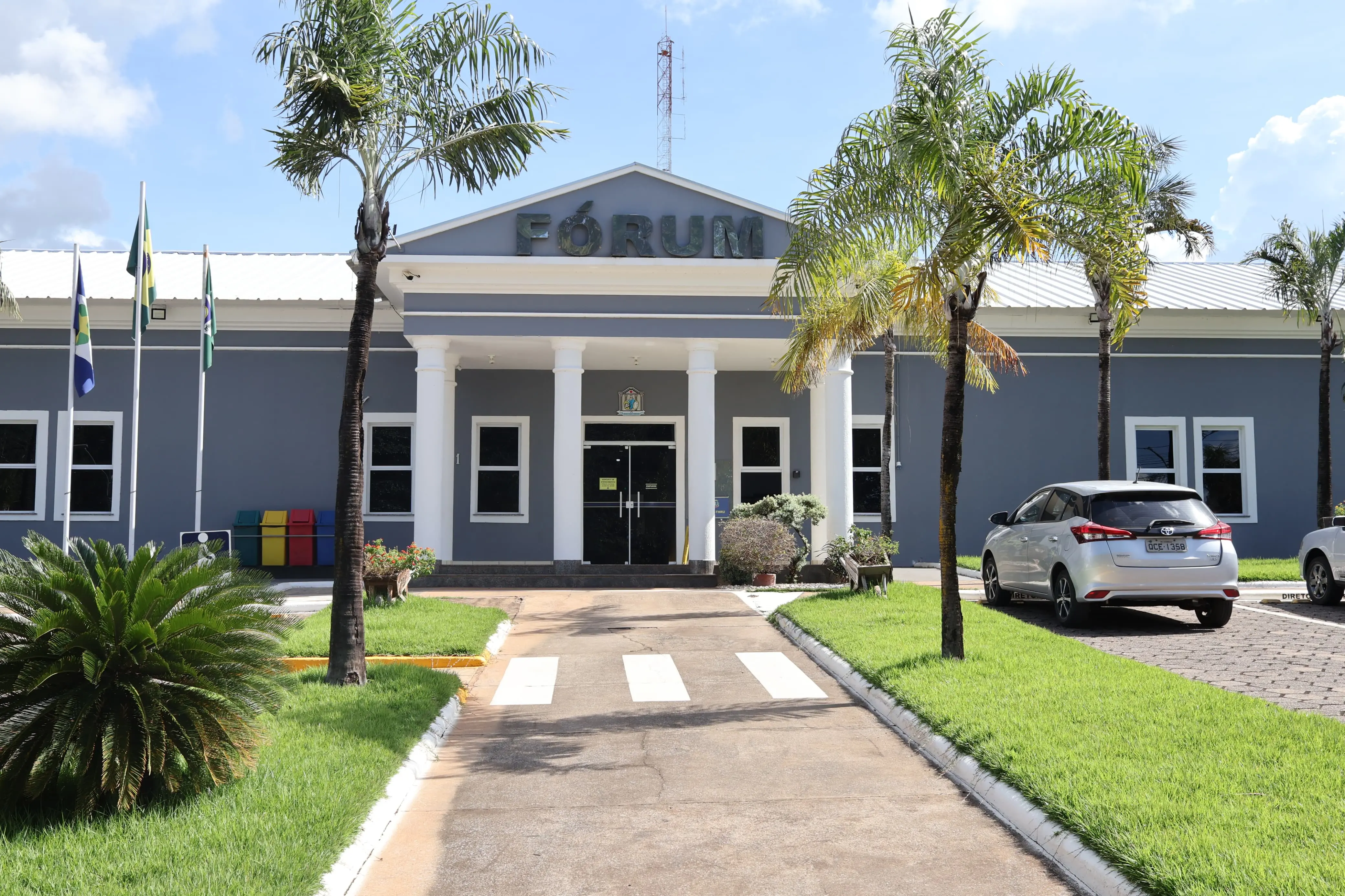 Fachada do Fórum da comarca, prédio térreo em tons de azul e branco, com colunas na entrada e a palavra “Fórum” no alto. Palmeiras ornamentam o jardim bem cuidado, com gramado, faixa de pedestres e vagas de estacionamento ao lado.