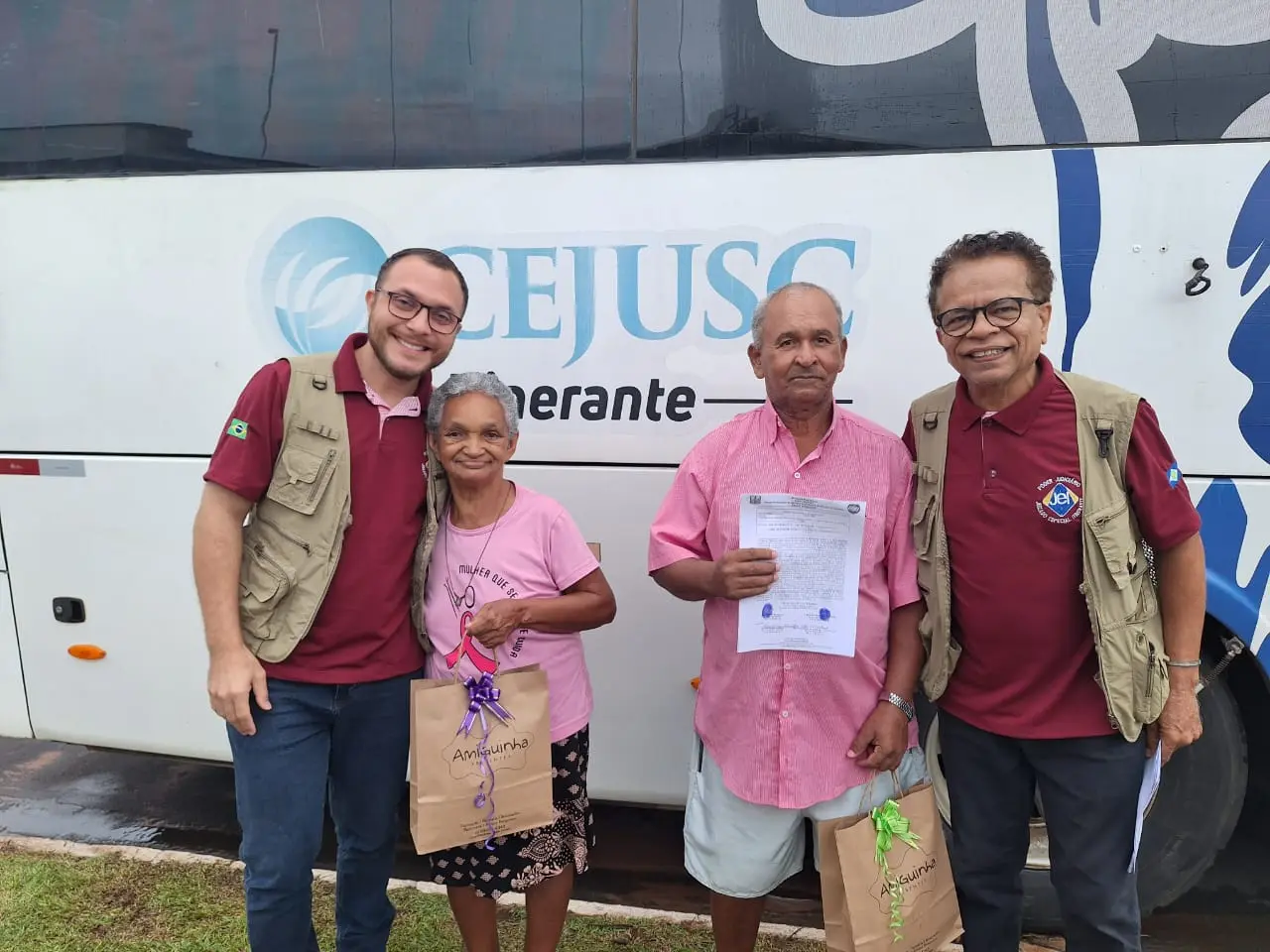 Quatro pessoas posam em frente ao ônibus do CEJUSC Itinerante. Dois servidores usam colete bege e camisa bordô. Um casal de idosos, sorridente, segura documentos e sacolas. Cena externa, clima de acolhimento e cidadania.