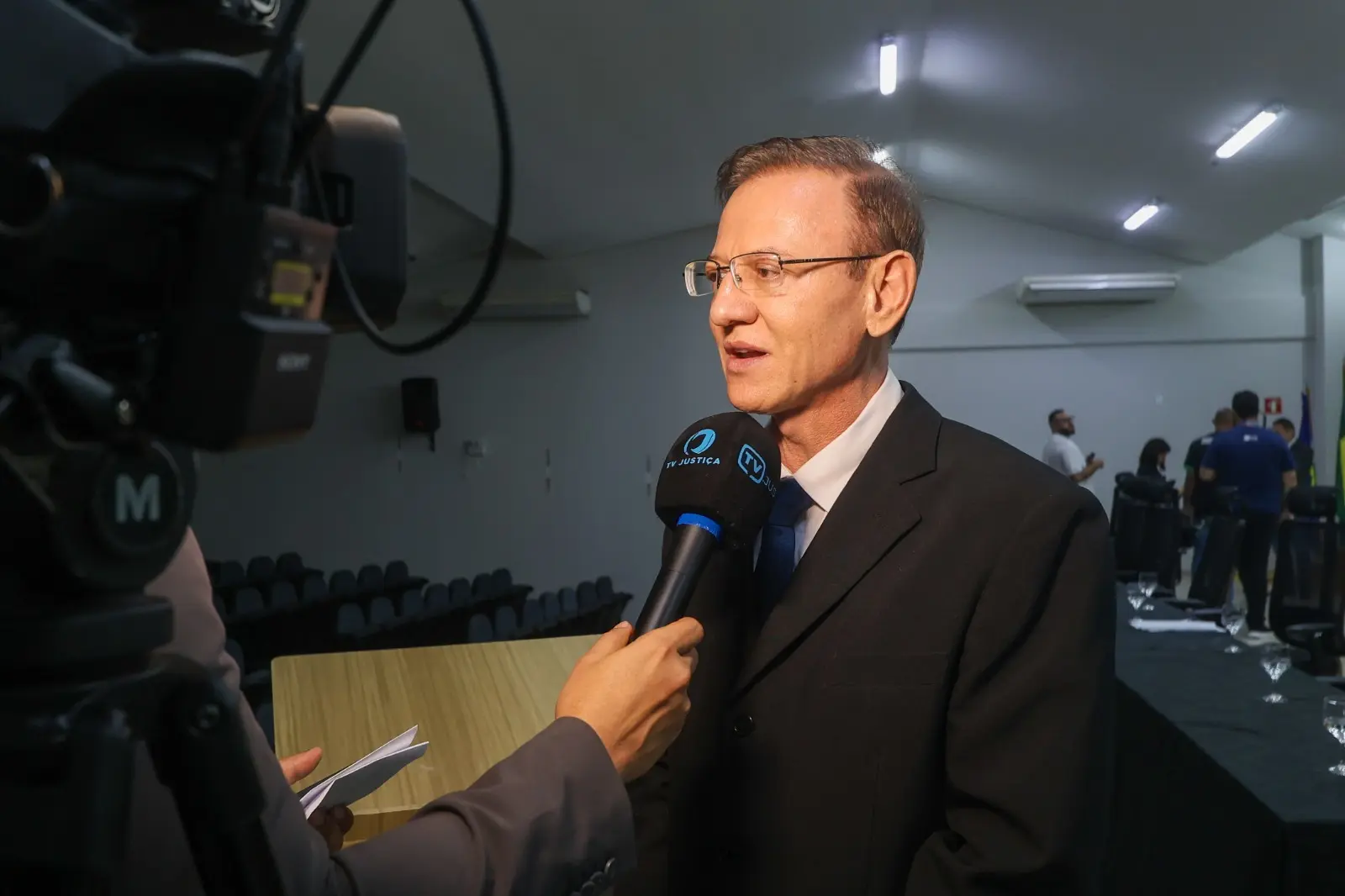 Homem adulto, de terno escuro, é entrevistado por equipe de comunicação em auditório ou sala de eventos. Ele fala ao microfone da TV Justiça, enquanto câmera grava a cena. Ao fundo, há cadeiras, mesa e outras pessoas no local, em ambiente formal.
