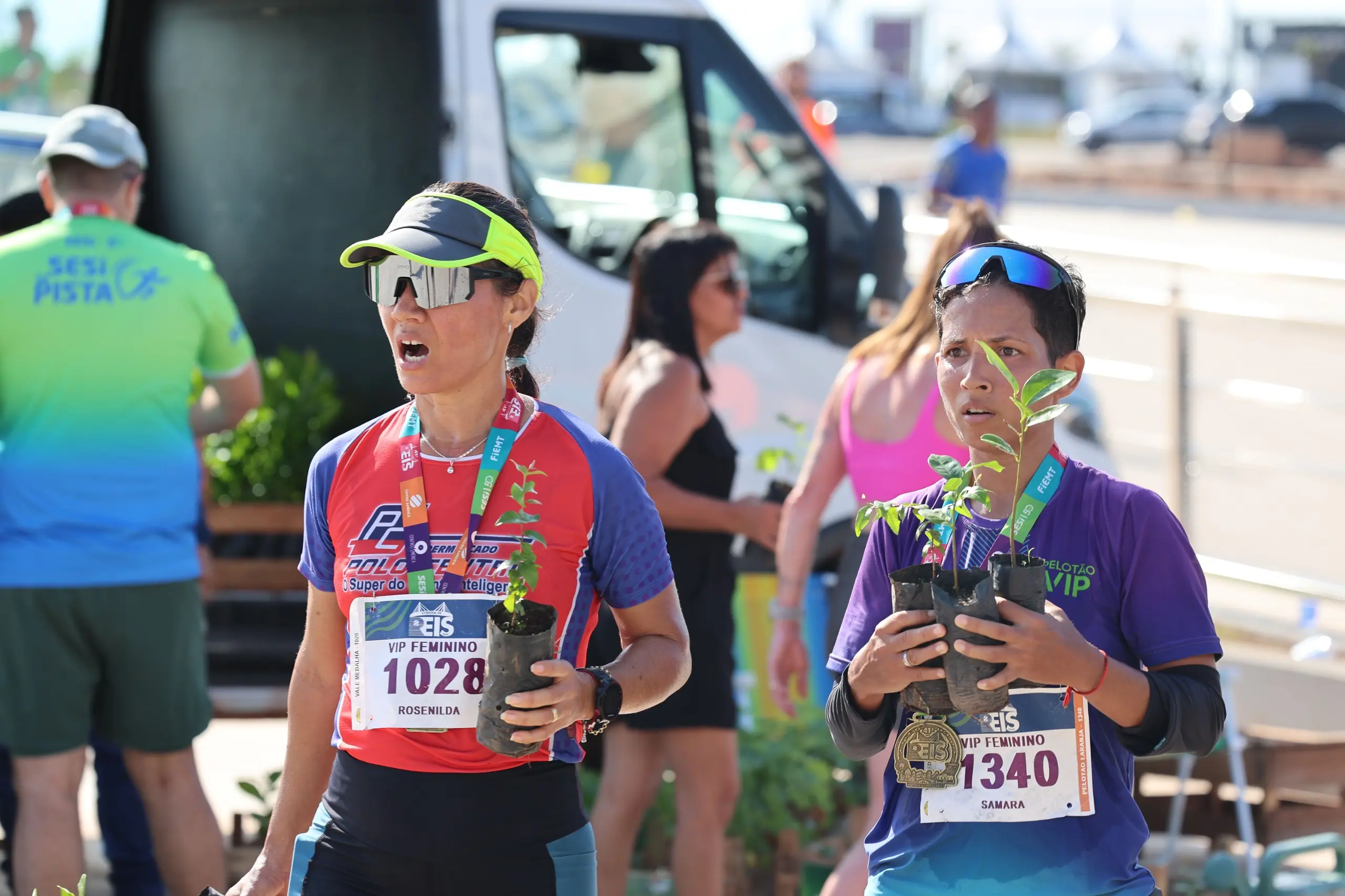 Duas mulheres usam camisetas esportivas, medalhas e números de peito, cada uma segurando mudas de árvores. A cena ocorre próxima a um veículo de apoio, com outros participantes passando atrás.