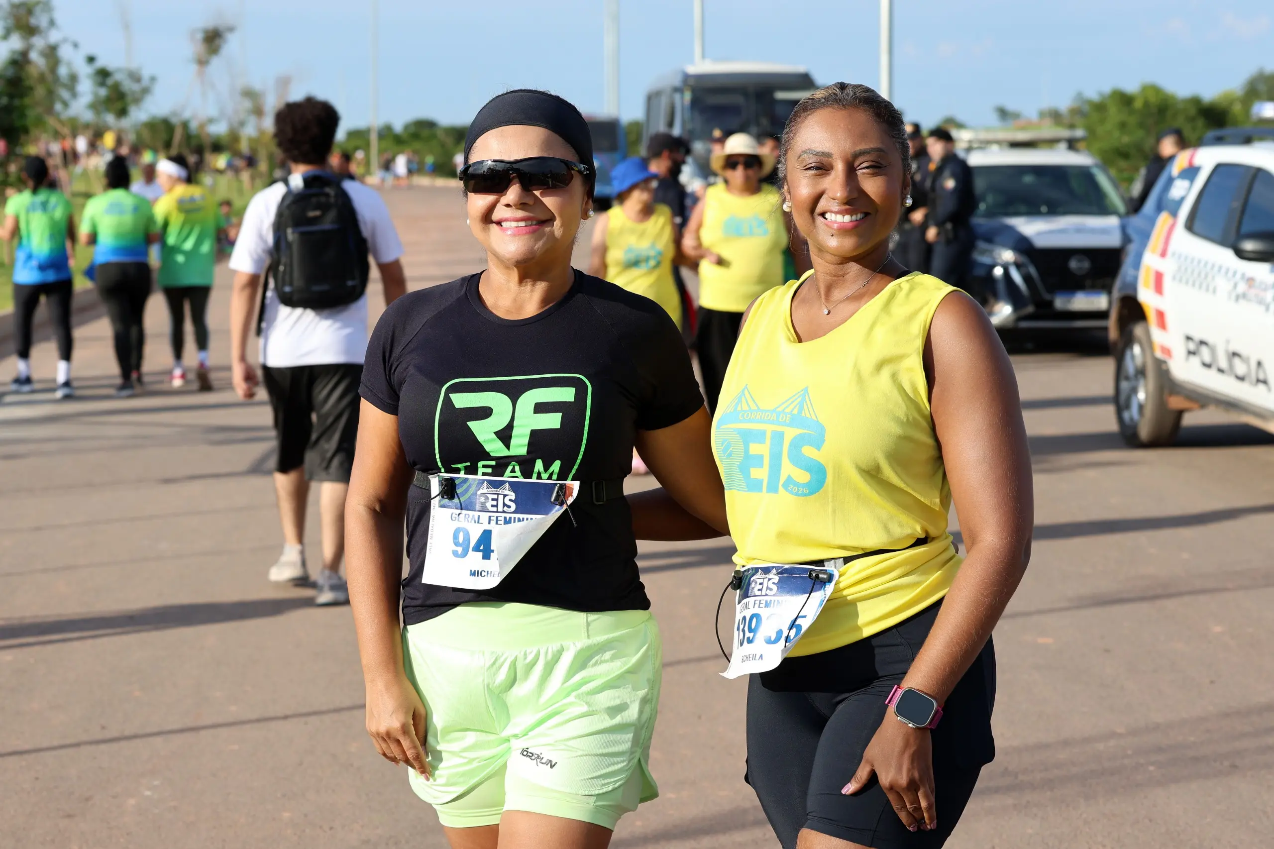 Duas corredoras caminham lado a lado após a corrida, usando roupas esportivas e medalhas. Ao fundo, outros atletas circulam pelo local de chegada.