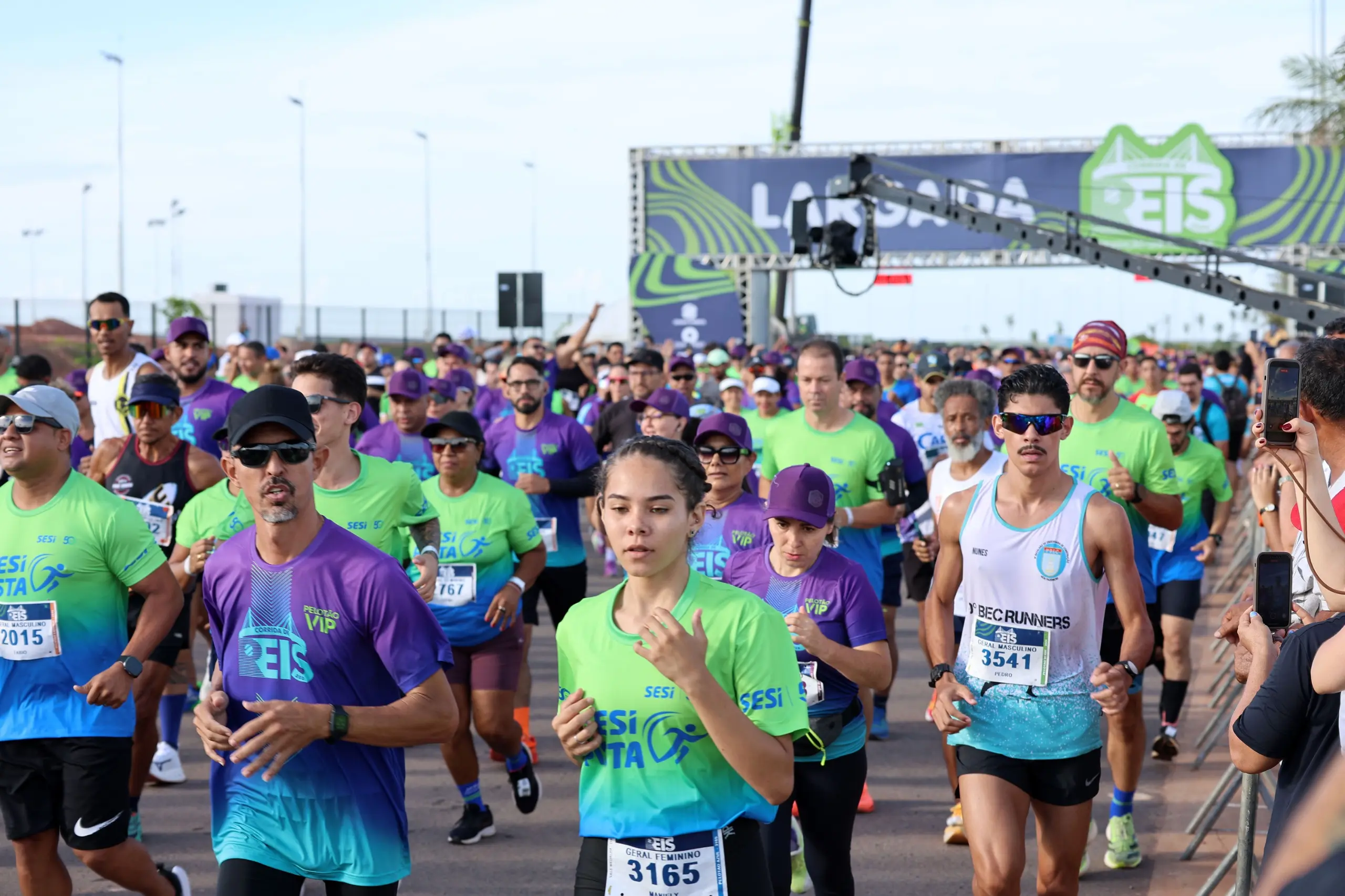 Vista ampla da largada mostra grande número de corredores distribuídos pela pista. O cenário inclui o pórtico oficial, cones e apoio logístico do evento esportivo.