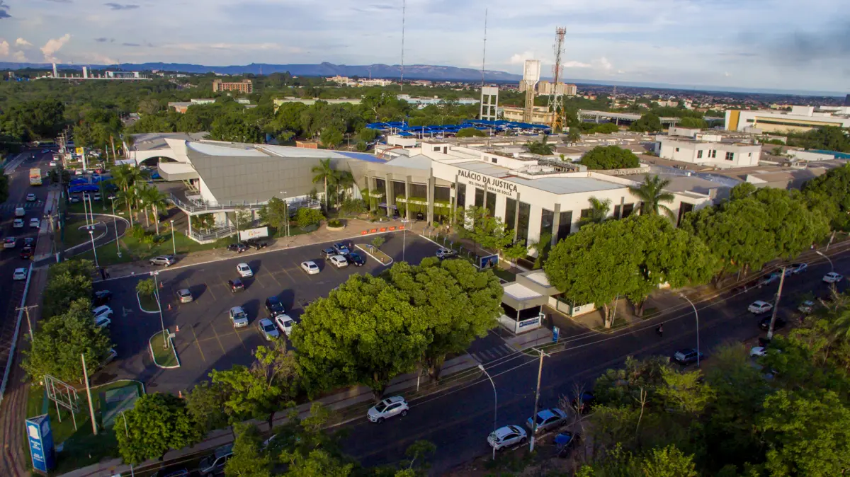 Vista aérea do Palácio da Justiça do TJMT, prédio moderno de fachada clara, cercado por árvores, estacionamento com veículos, vias asfaltadas e paisagem urbana ao fundo, sob céu parcialmente nublado.