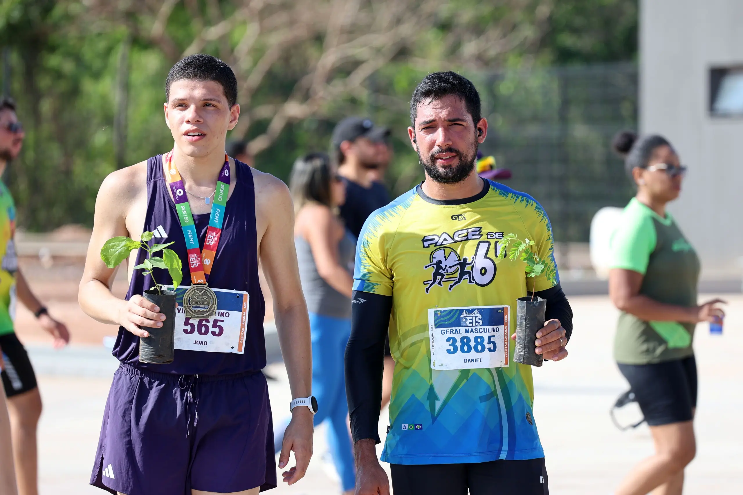 Dois corredores caminham lado a lado após a prova, ambos com medalhas no pescoço e mudas de árvores nas mãos. Eles estão em espaço aberto, com outros atletas dispersos e vegetação ao fundo.