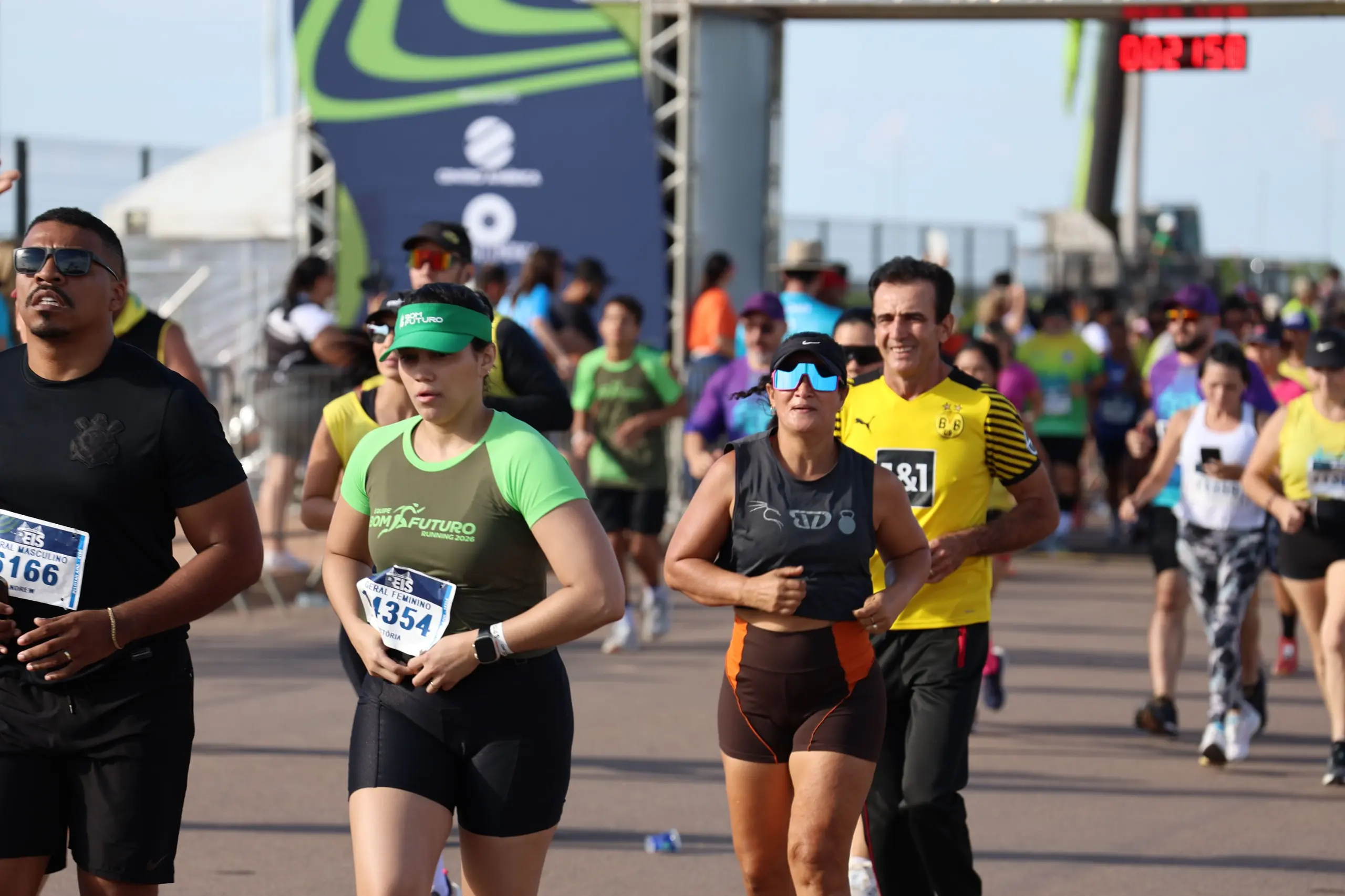 Corredores seguem juntos durante a prova, ocupando a pista em grupo compacto. Pessoas usam camisetas coloridas, bonés e óculos, em ambiente aberto e ensolarado.