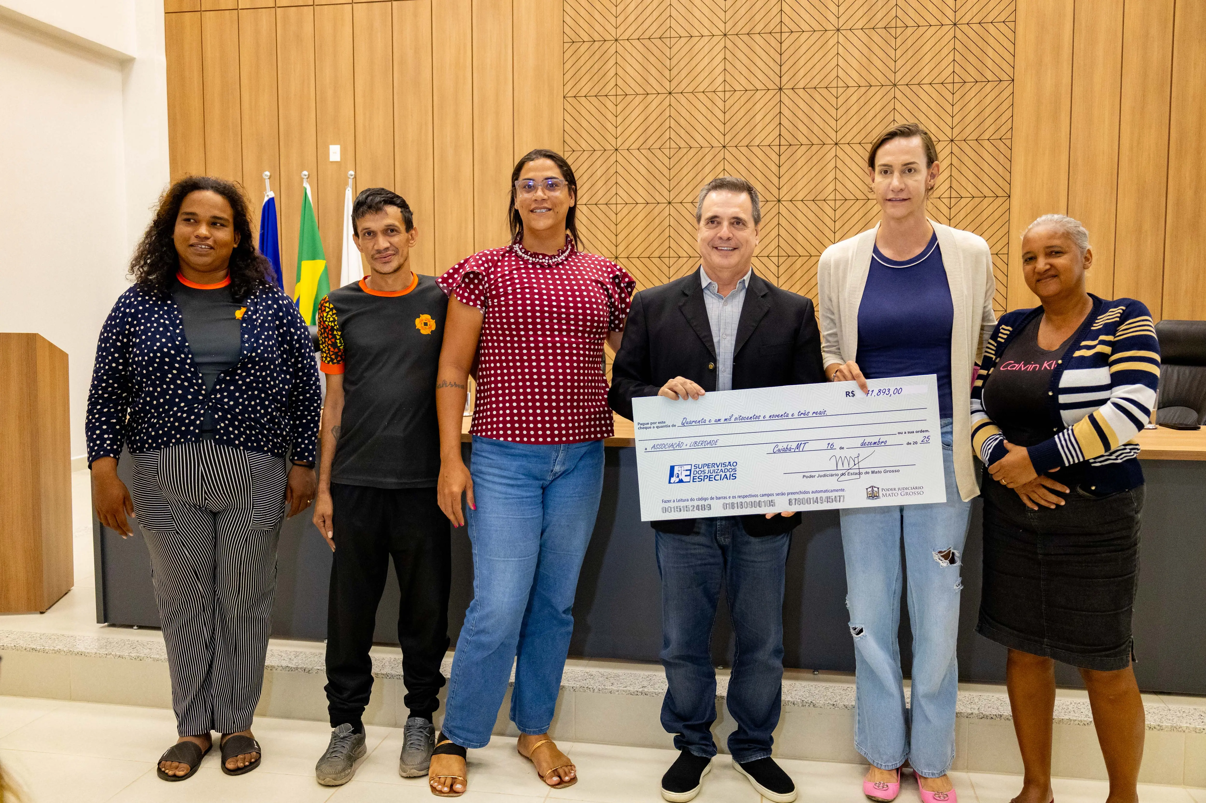 Pessoas reunidas no plenário exibem cheque simbólico em ação de entrega institucional. Todos estão em trajes casuais e formais, com bandeiras e estrutura oficial do Tribunal ao fundo.