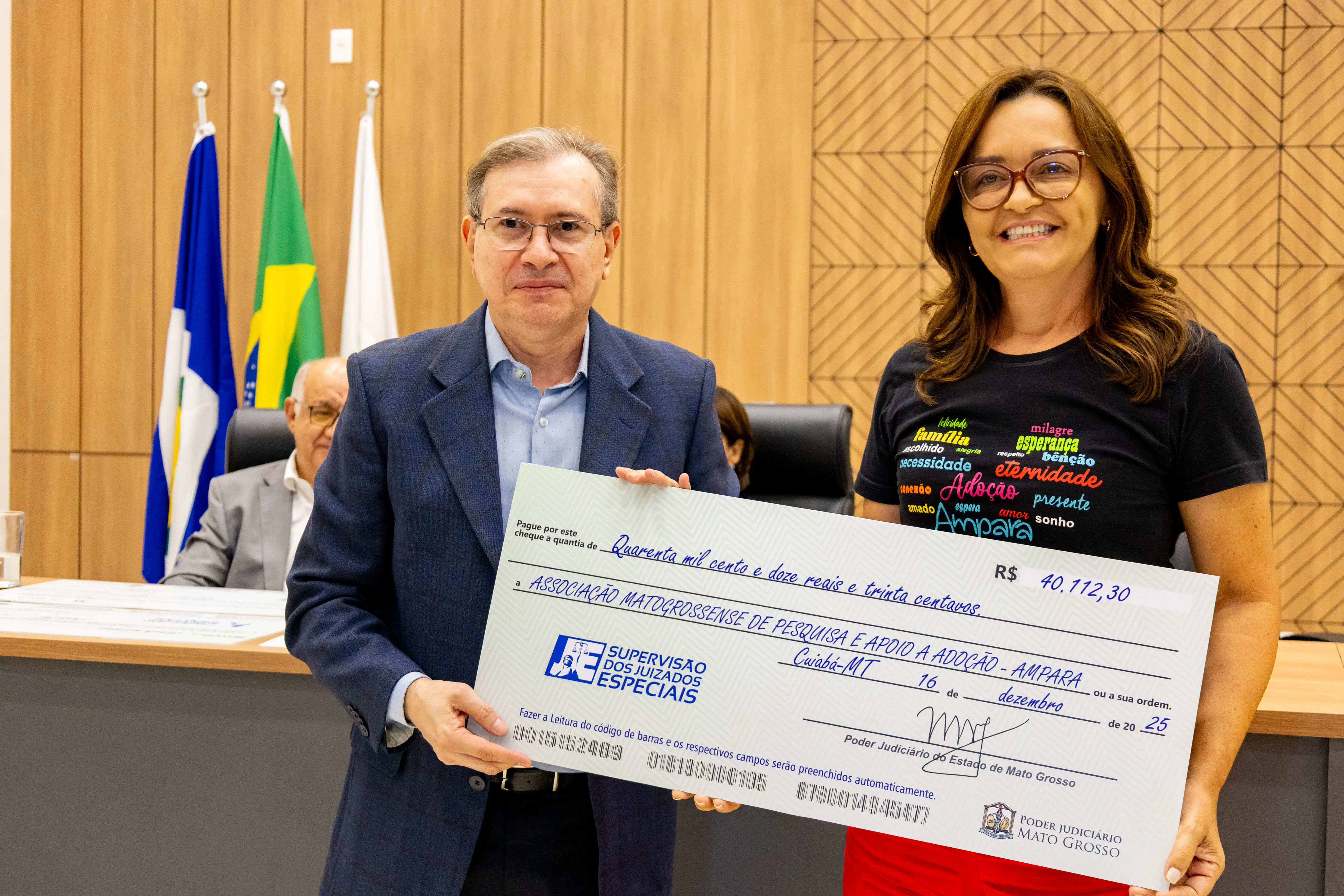 Homem e mulher posam à frente de mesa institucional segurando cheque simbólico. Ambos sorriem para a câmera. Ao fundo, bandeiras oficiais e autoridades sentadas, indicando ato formal de entrega de recursos.