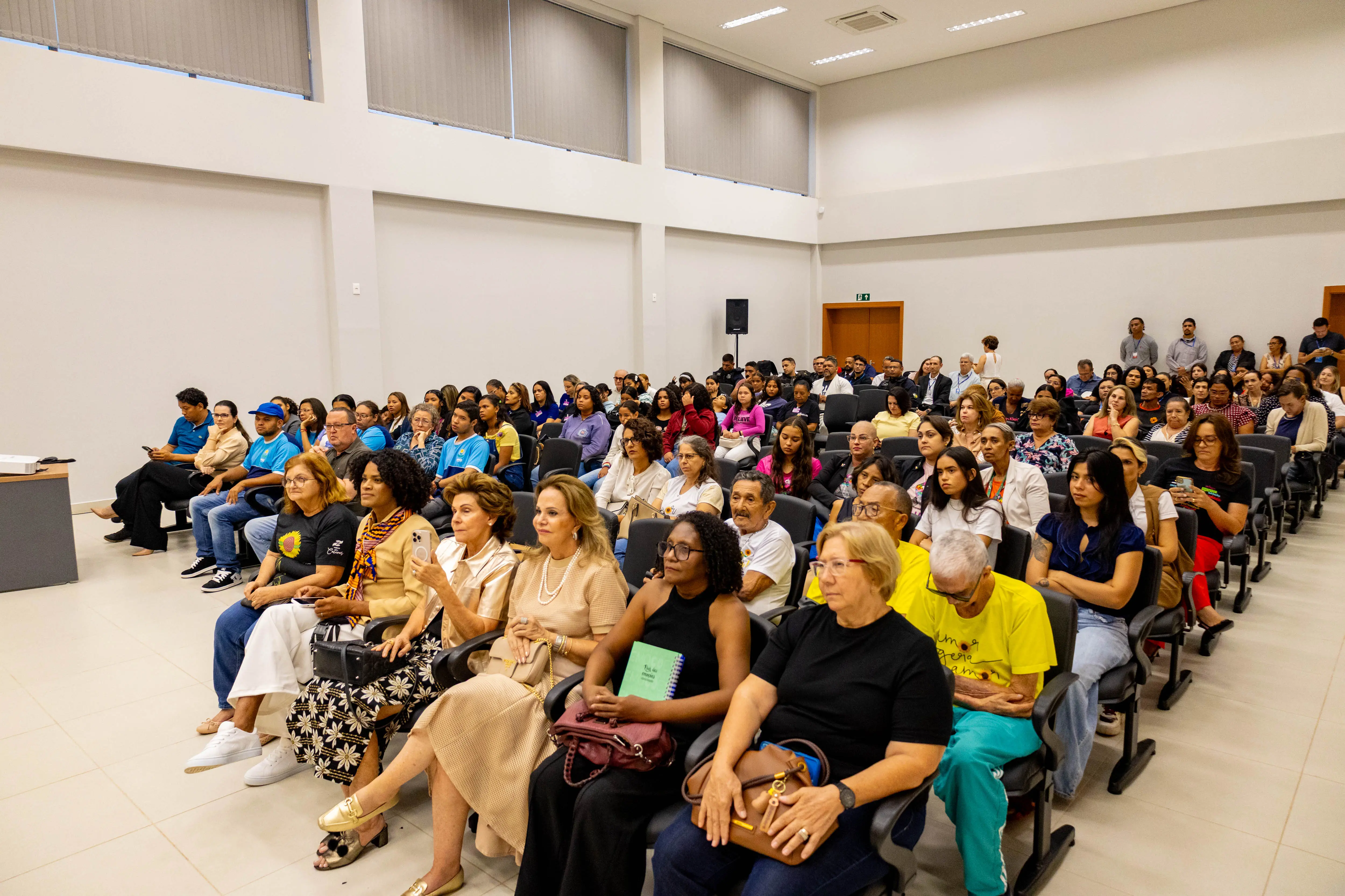 Auditório amplo com dezenas de pessoas sentadas, de diferentes idades e perfis, atentas à programação. As cadeiras são pretas, o espaço é bem iluminado e transmite clima de participação comunitária e interesse coletivo.