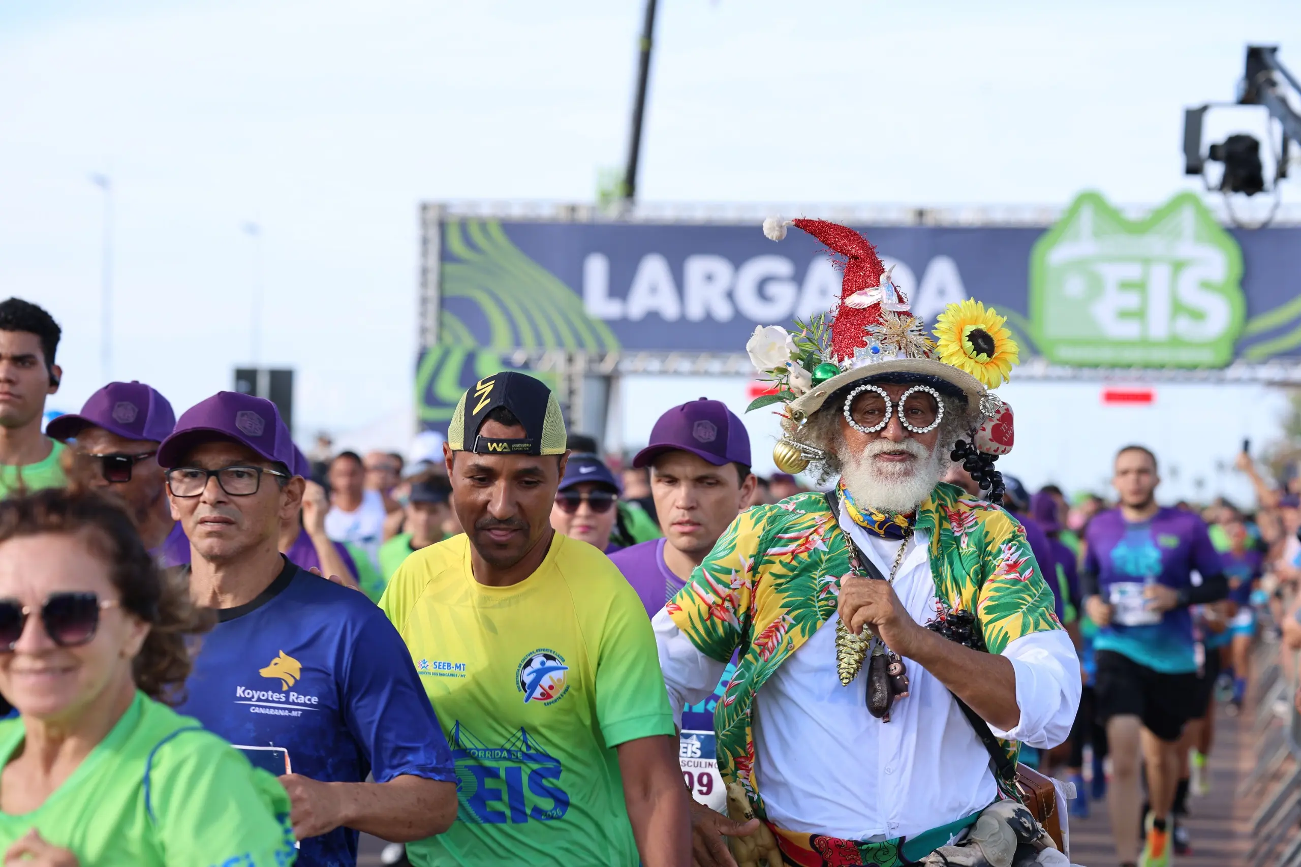 Grupo diverso de participantes passa pelo pórtico de largada, alguns sorrindo e outros concentrados. O local é amplo, com bandeiras, grades e apoio organizacional ao redor.