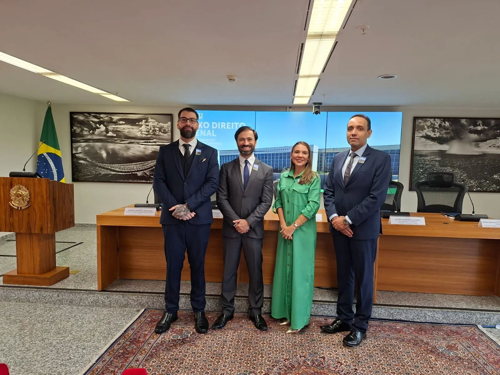  Quatro pessoas (três homens de terno e uma mulher de vestido longo verde) posam em pé em um auditório jurídico. Ao fundo, uma bancada de madeira, fotos de paisagens e o logotipo do STJ em um telão. O clima é formal e institucional.