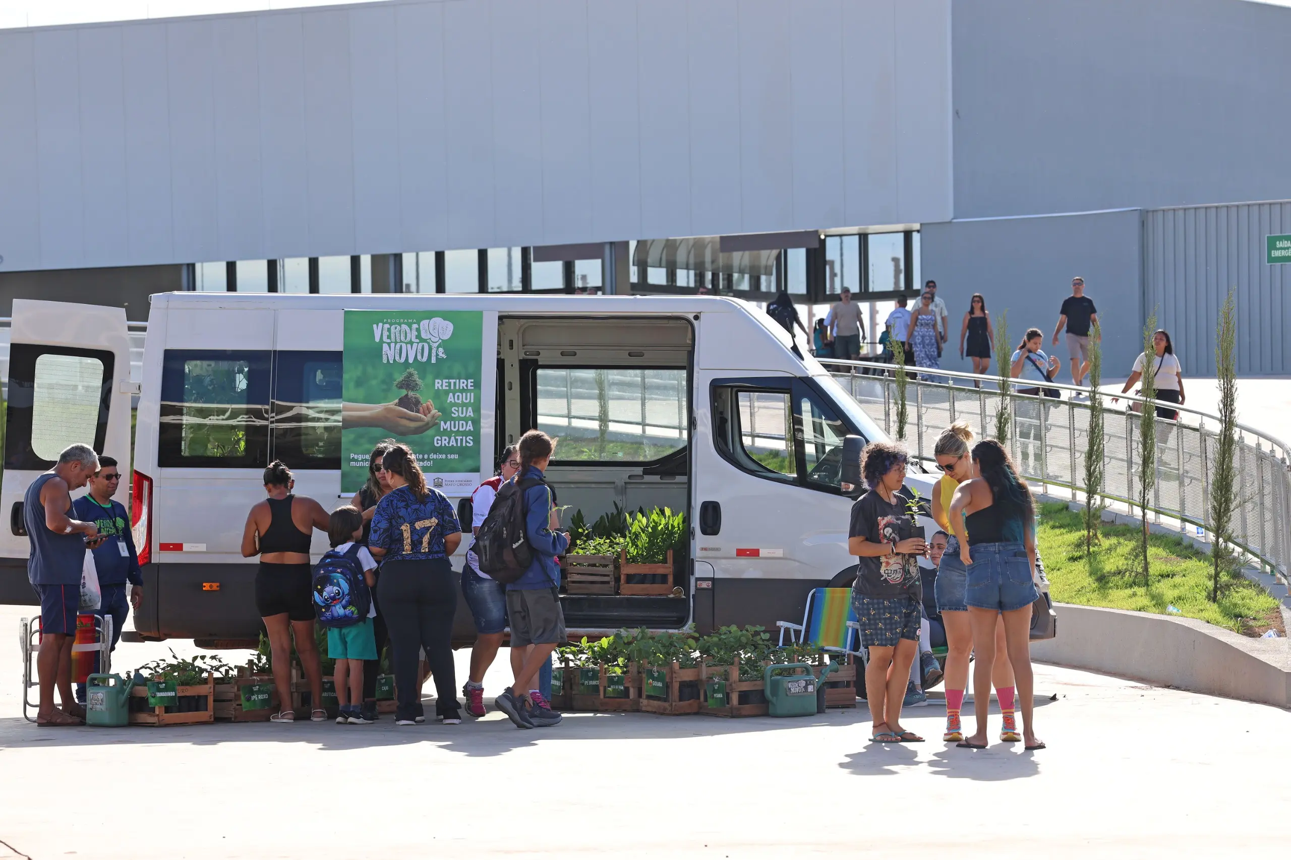 Área externa do evento com van do projeto Verde Novo estacionada. Participantes da corrida se aproximam para retirada de mudas, enquanto equipe organiza caixas de plantas em espaço amplo e ensolarado.