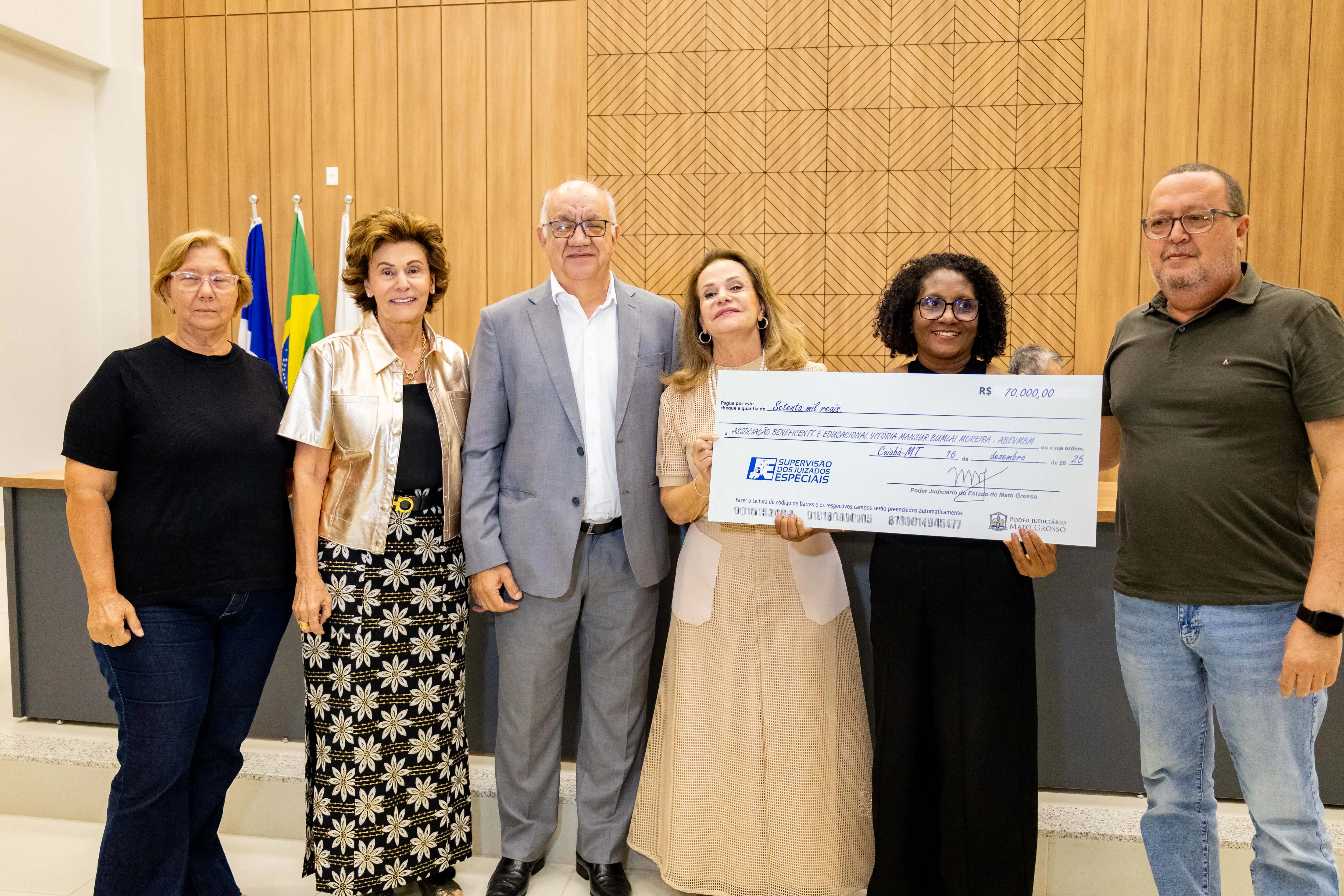 Quatro mulheres e um homem posam com cheque simbólico. A mulher de cabelos curtos aparece novamente ao centro. O grupo sorri, em ambiente institucional que reforça parceria e apoio a projetos sociais.