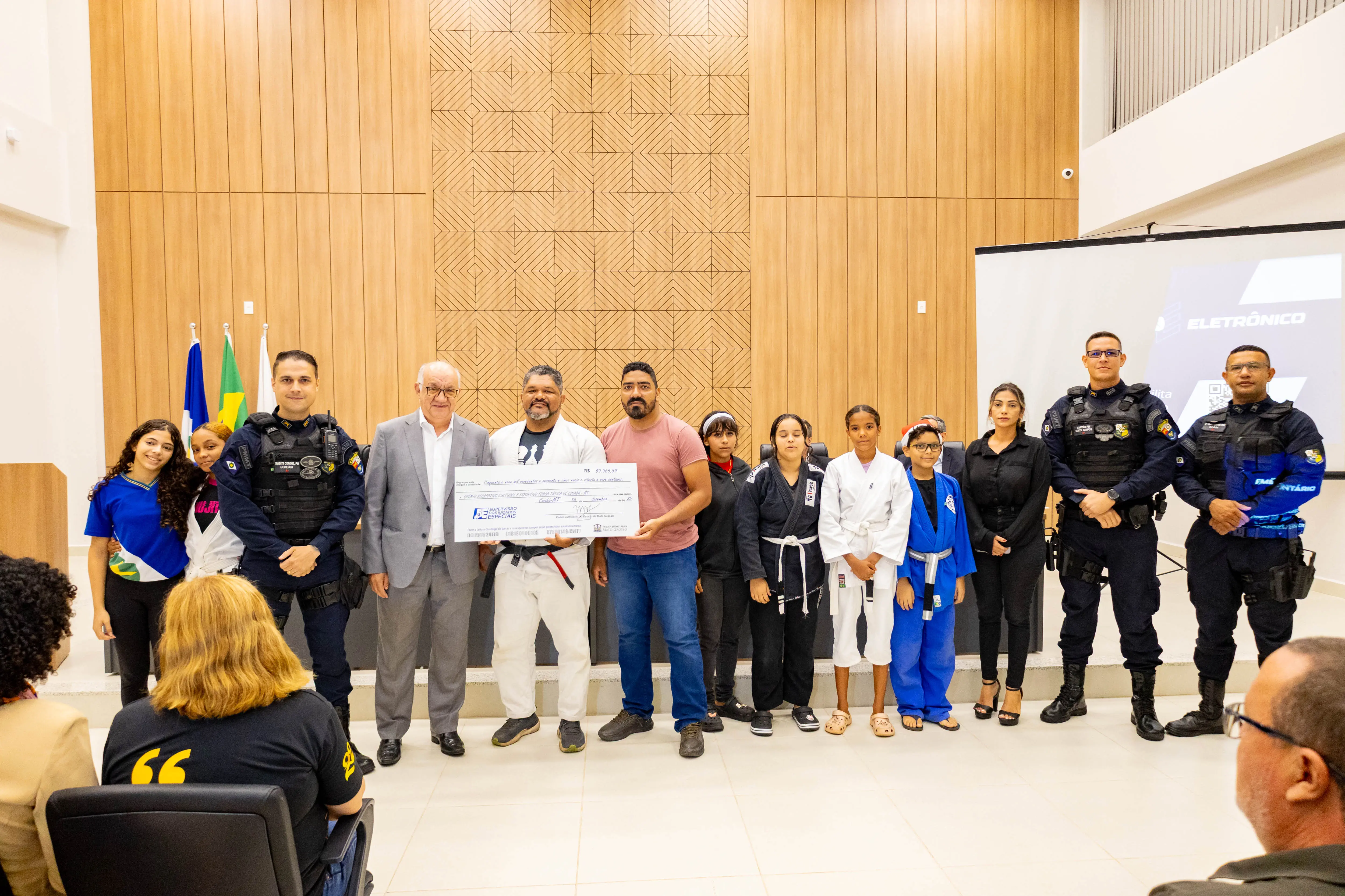Grupo com policiais fardados, crianças e adultos posam juntos segurando cheque simbólico. A diversidade de participantes destaca ação social integrada, em espaço oficial do Judiciário, com público acompanhando.