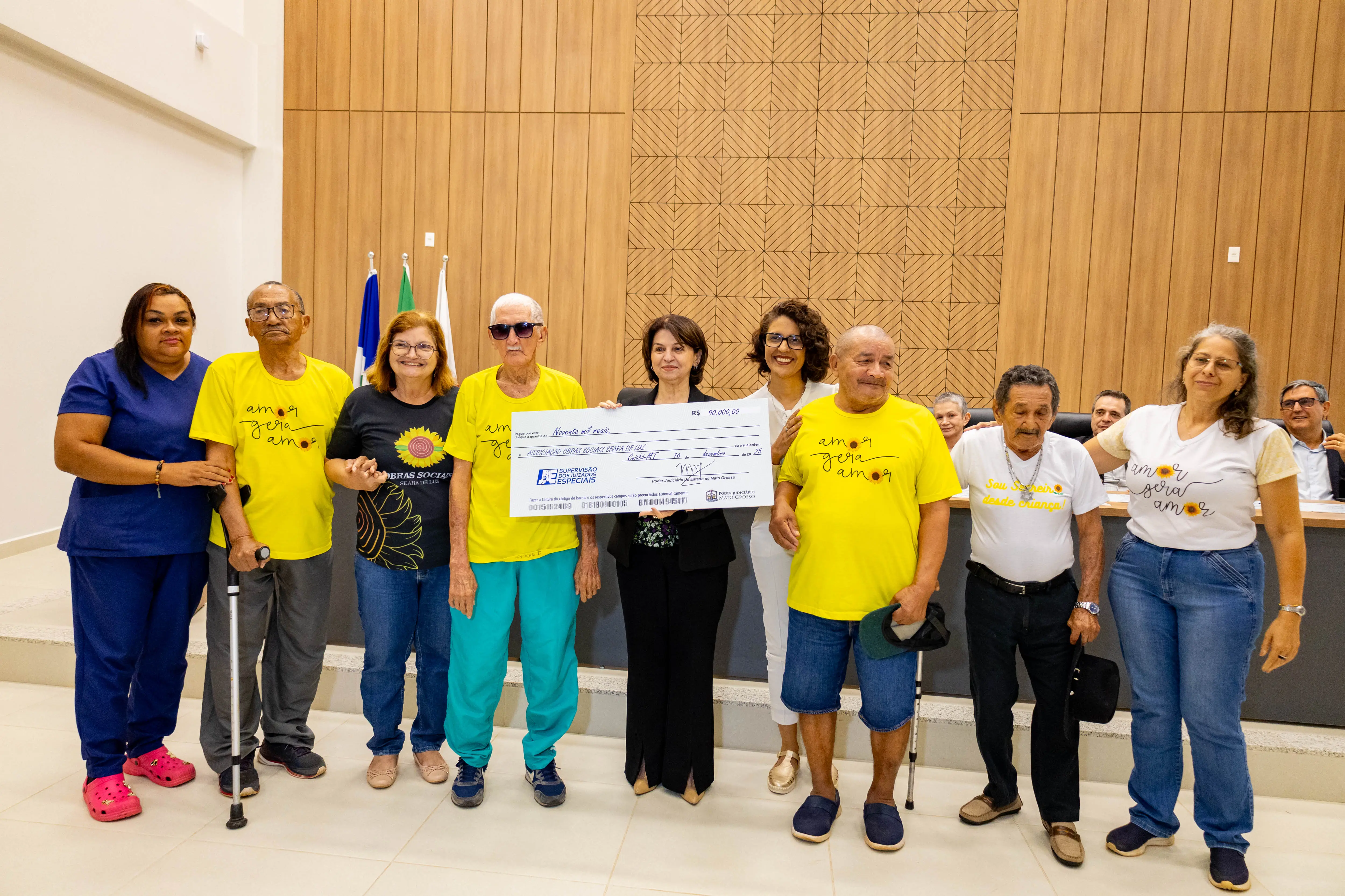 Grupo diverso de homens e mulheres, alguns idosos, posam lado a lado segurando cheque simbólico. Muitos usam camisetas coloridas. A cena ocorre em auditório oficial, marcando momento de reconhecimento e apoio social.