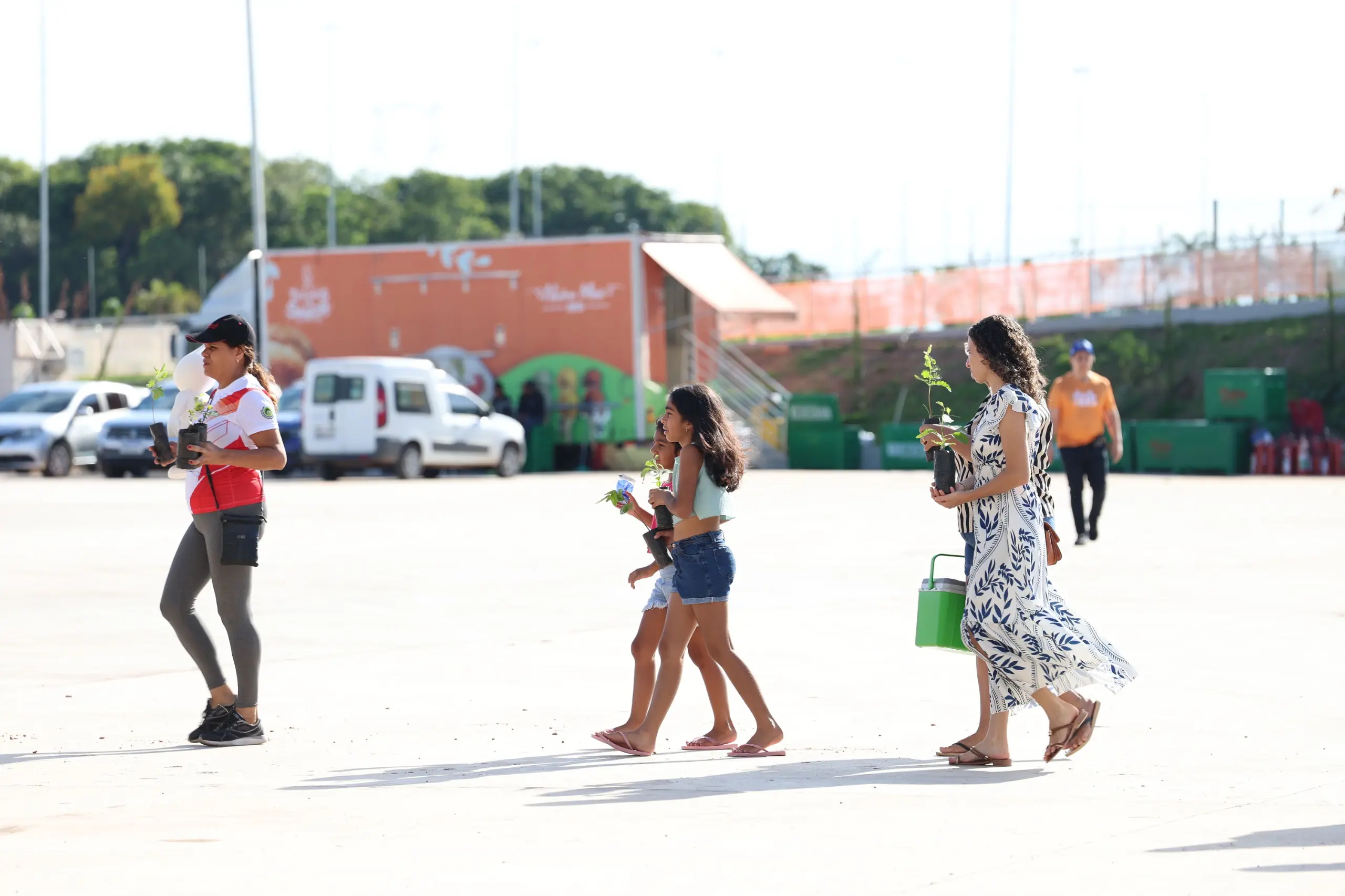 Pessoas c aminham com mudas de plantas nas mãos. O ambiente é amplo e cinza