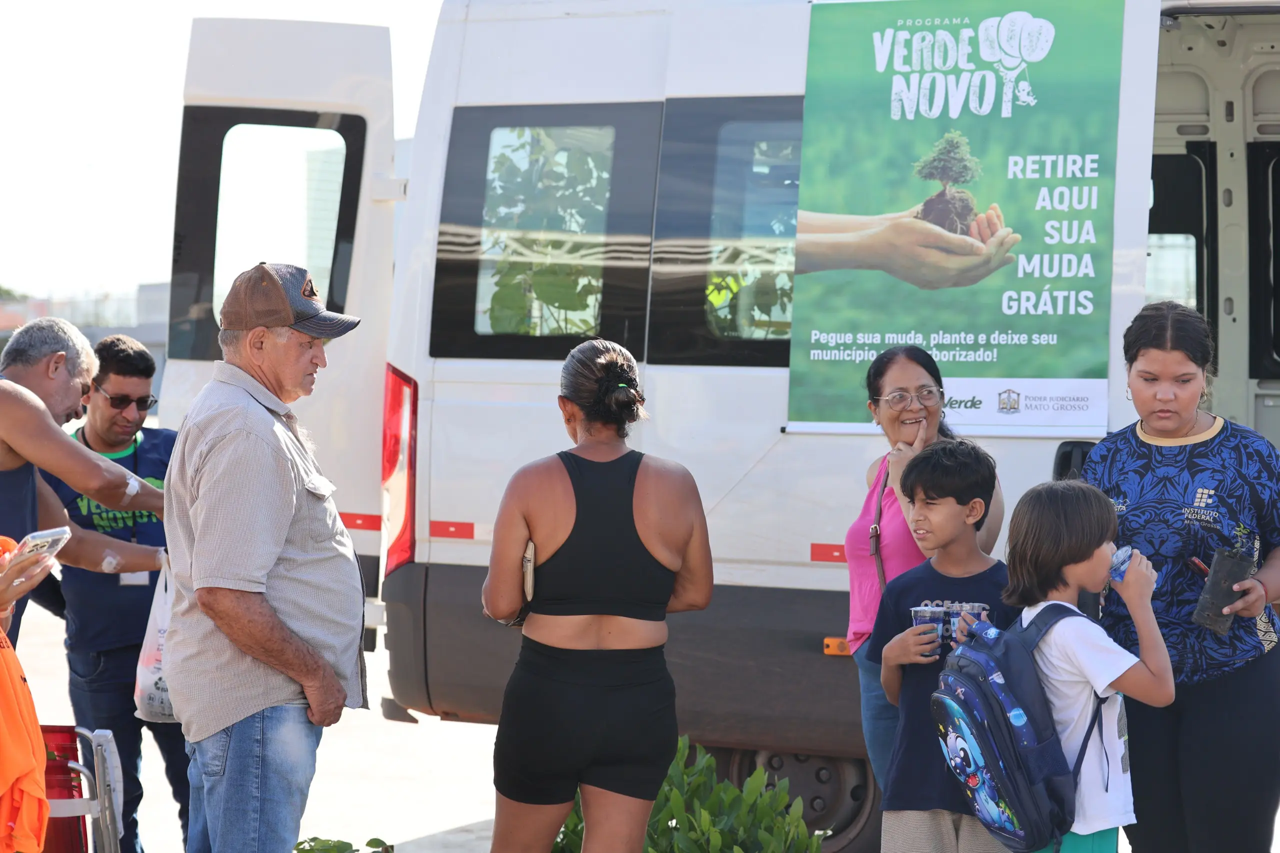 Mulheres e crianças se reúnem em frente à van do projeto ambiental. Adultos conversam enquanto os pequenos observam e seguram mudas, em ambiente aberto após a corrida.