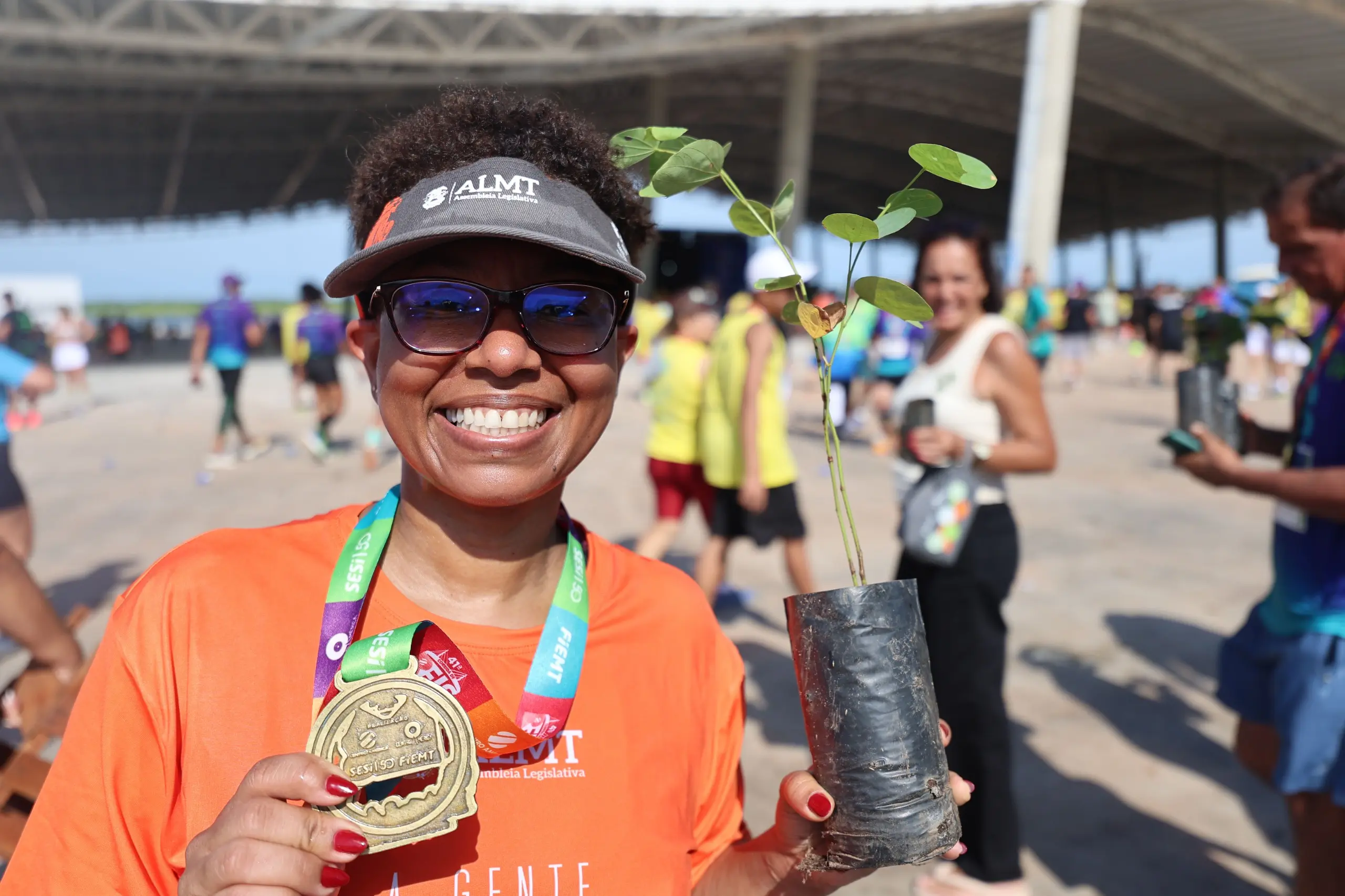 Mulher sorridente, usando camiseta laranja, viseira e óculos escuros, exibe medalha da corrida e segura uma muda de árvore em saco plástico. A foto foi feita em área aberta do evento, com participantes e pavilhão ao fundo.