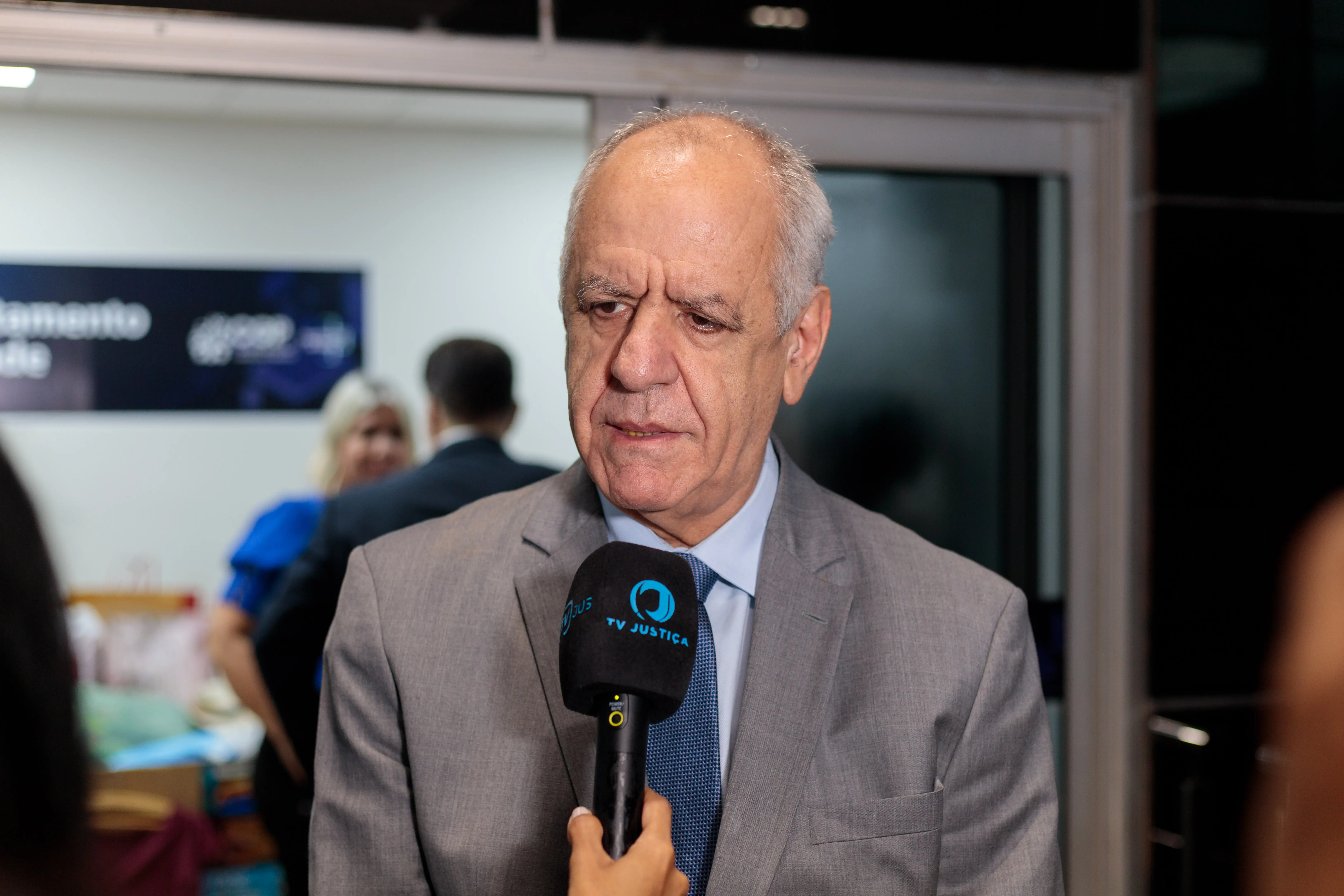 Ussiel Tavares concede entrevista duran te entrega dos itens para doação á Casa de Amparo. Ele é um homem branco, cabelos grisalhos, usa blaser cinza, gravata azul e camisa azul clara