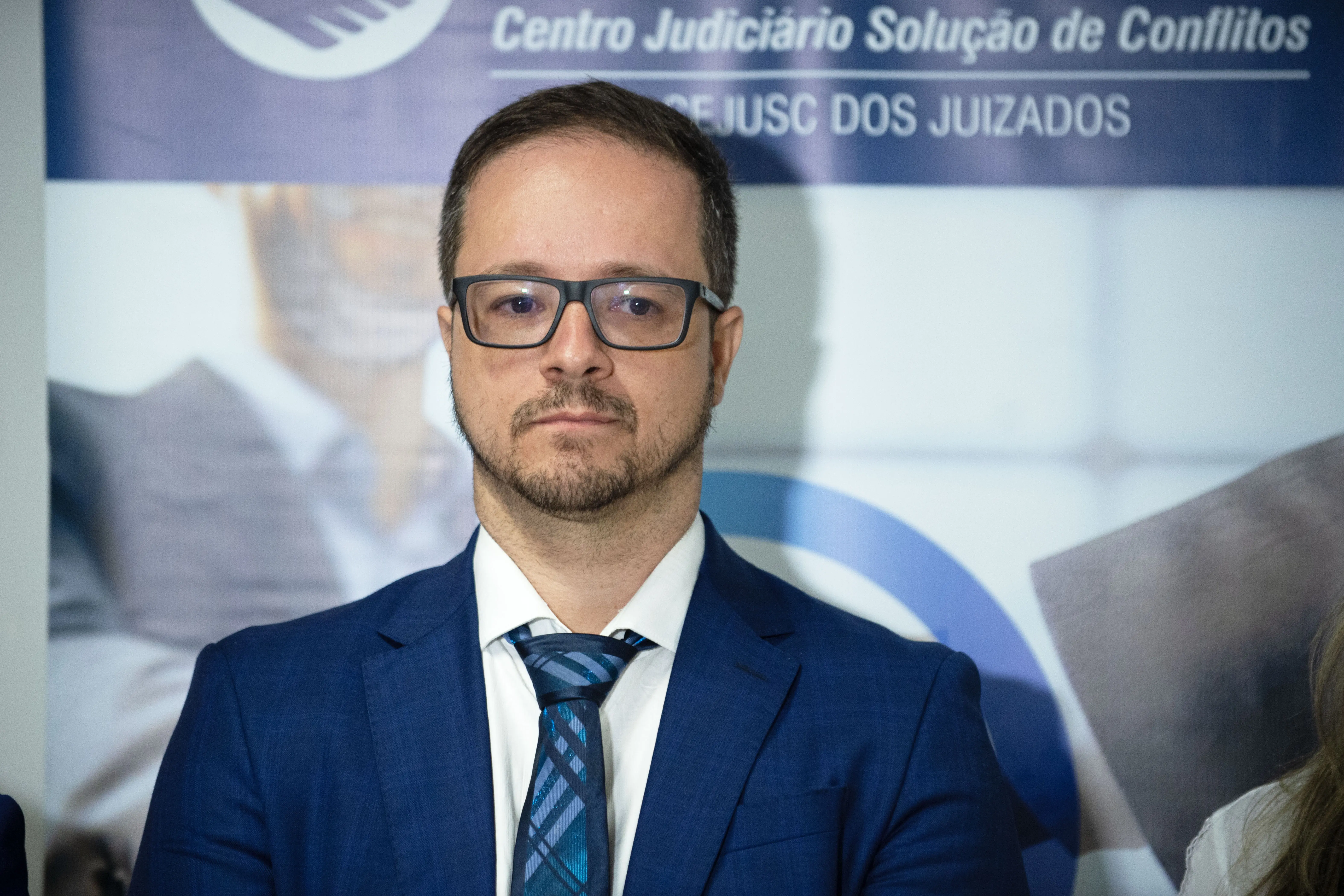 Promotor Alberto Biazotto Júnior. Ele é um homem branco, com cavanhaque, cabelo curto castanho. Usa óculos de grau com armação escura, camisa branca, blaser e gravata azuis