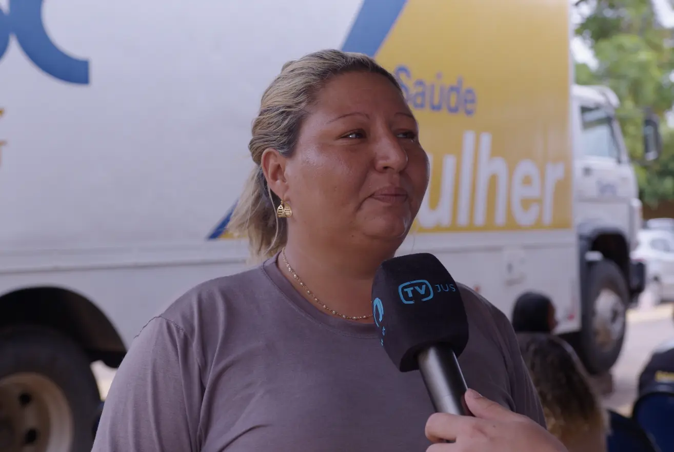 Márcia Barros durante entrevista à TV.Jus, em frente à carreta Sesc Saúde Mulher. ela é uma mulher de 42 anos, parda, com cabelo loiro preso, usando camiseta cinza e brinco dourado.