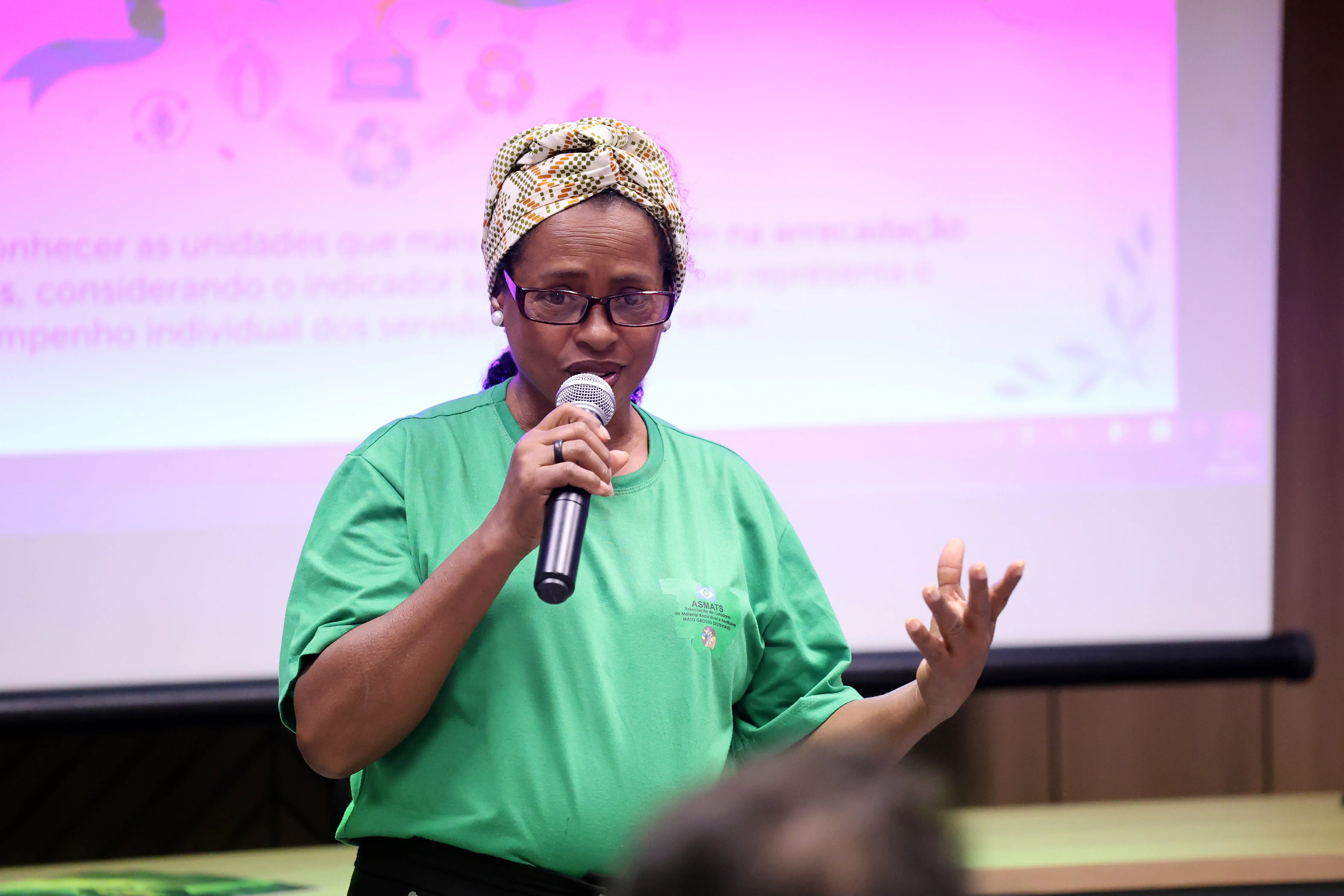 Mulher com camiseta verde e turbante fala ao público segurando microfone. Ao fundo, projeção rosa com informações do ReciclaJud. Ela gesticula enquanto apresenta. Local é sala de treinamento do TJMT, com luz interna e participantes sentados.