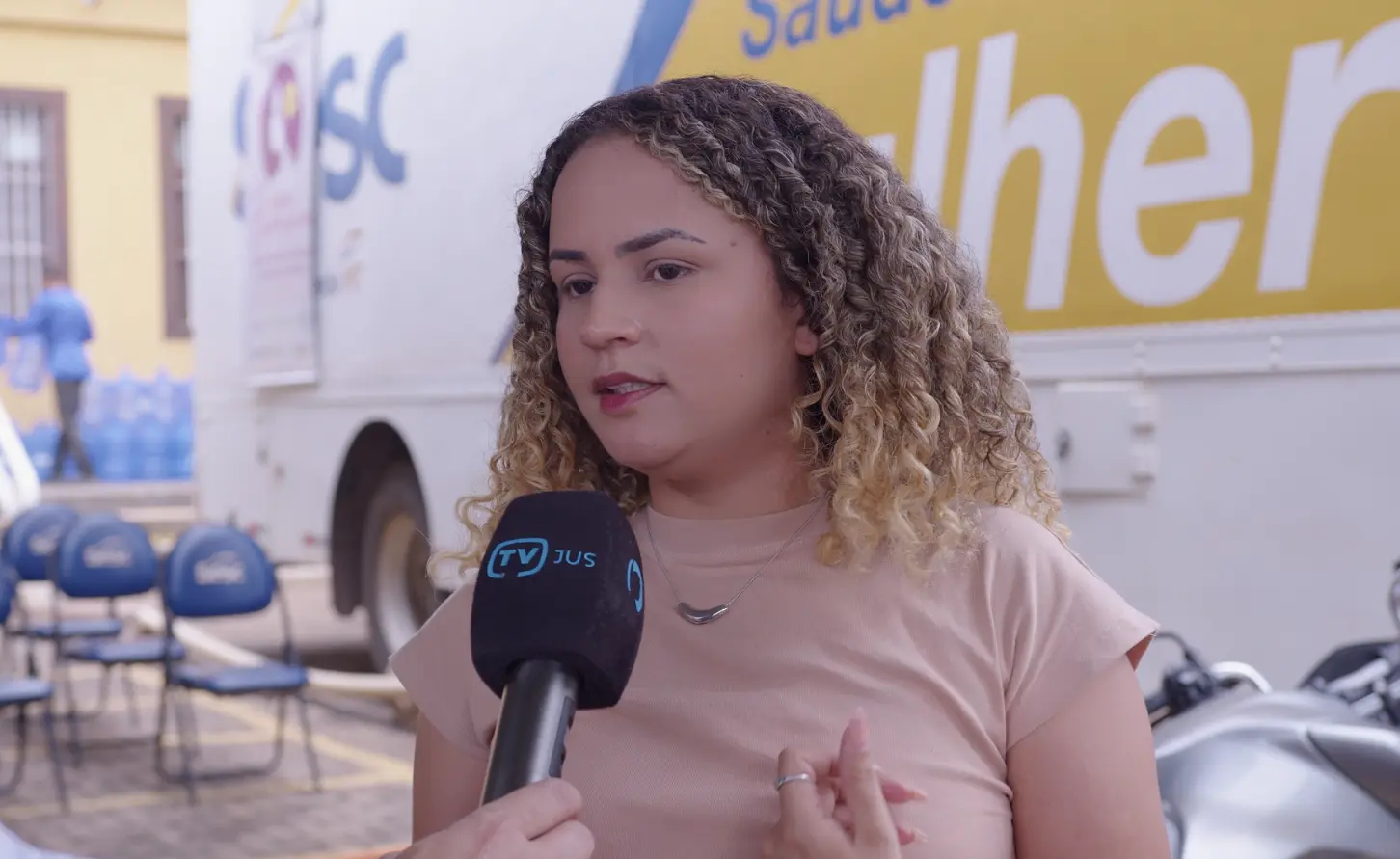 Ana Vitória Lustosa durante entrevista à TV.Jus, em frente à carreta Sesc Saúde Mulher. Ela é uma jovem de pele clara, olhos castanhos, cabelos cacheados e com mechas loiras, usando blusa bege.