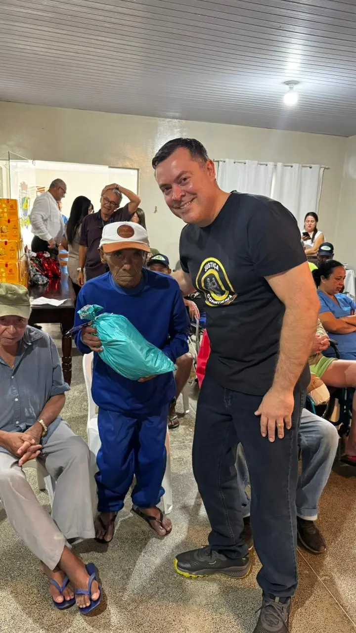 Juiz Leonísio Salles com camiseta da Justiça Comunitária entrega um presente azul a um idoso de boné branco. Ambos sorriem timidamente em um salão cheio de cadeiras e outros participantes da ação social.
