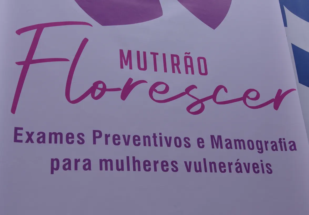 Detalhe do banner do Mutirão Florescer pendurado. O banner é rosa e lilás, onde se lê:  Mutirão Florescer - exames preventivos e mamografia para mulheres vulneráveis.