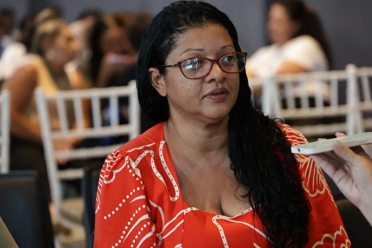 A mulher usa vestido vermelho estampado e óculos, com cabelos longos e úmidos sob um lado do ombro. Ela olha atentamente para quem a entrevista. Ao fundo, pessoas sentadas indicam ambiente de evento ou palestra.