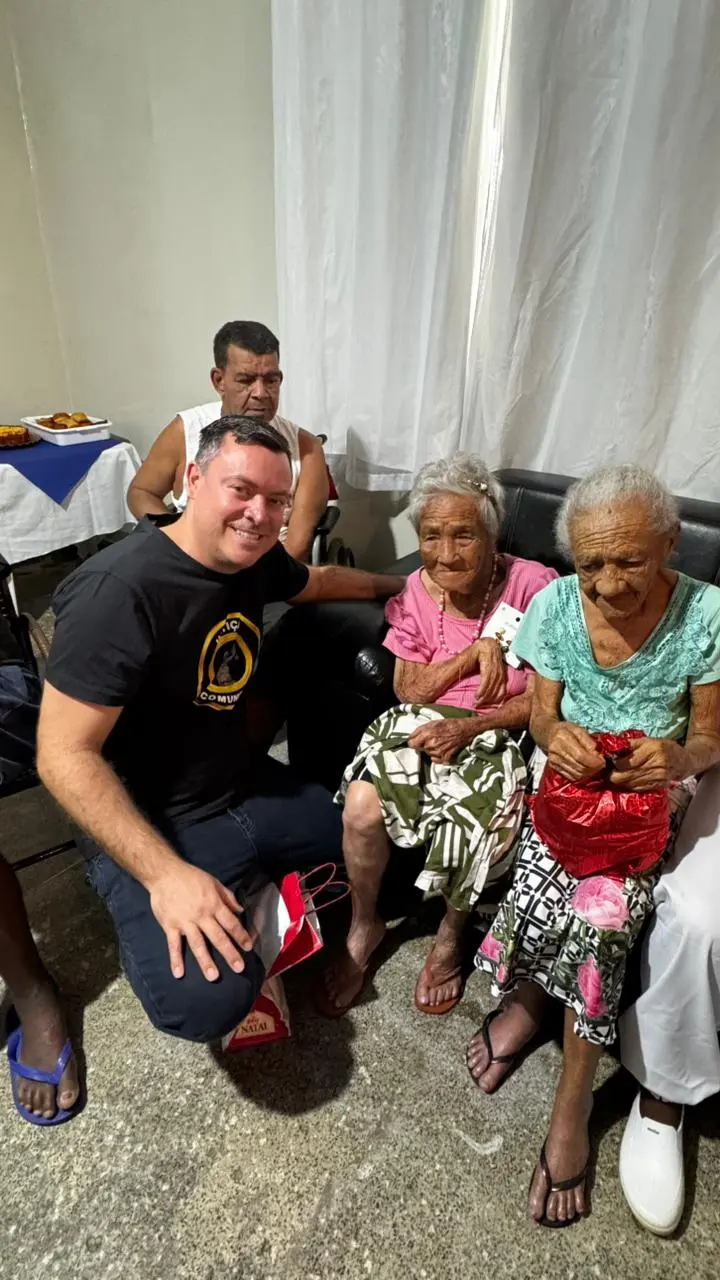 Juiz Leonísio Salles posa ajoelhado ao lado de duas idosas sentadas, que seguram presentes vermelhos. O ambiente é simples, com cortinas brancas e cadeiras. A ação reúne idosos em clima afetuoso.