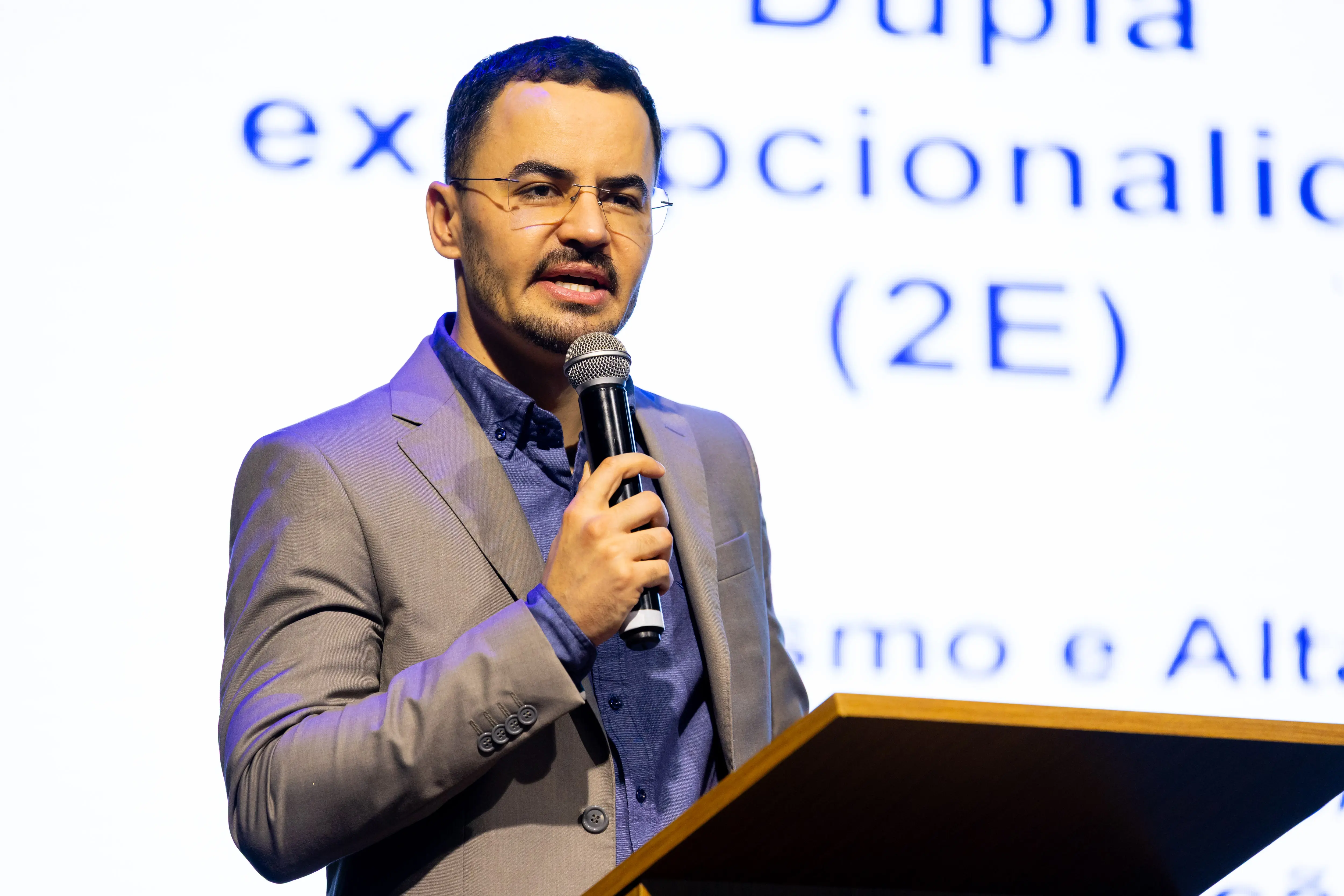 Em close, o palestrante segura o microfone enquanto discursa diante de tela branca com texto projetado. Ele usa terno cinza e camisa azul, iluminado por luz de palco que destaca sua expressão concentrada e o ambiente técnico do evento.