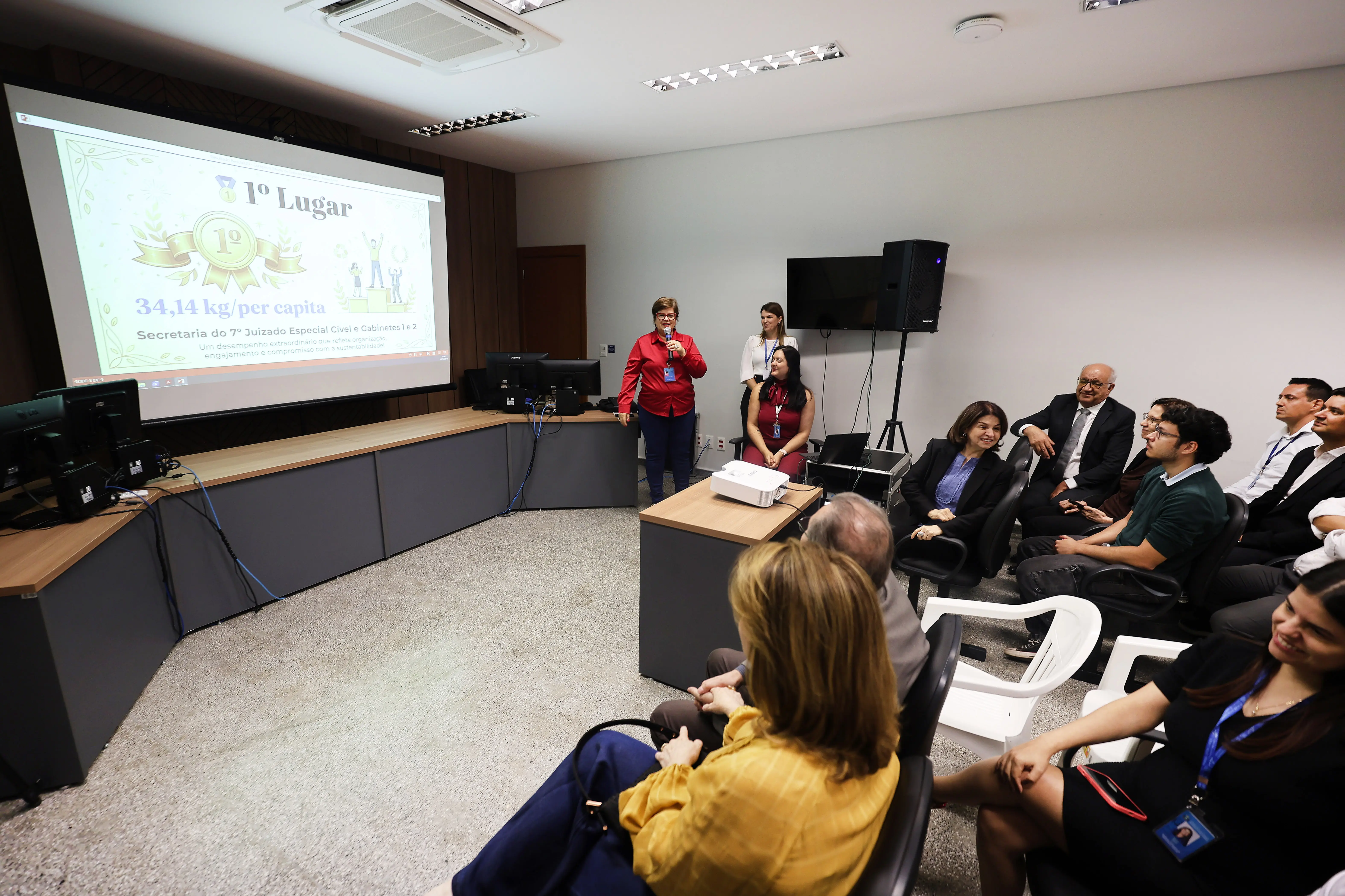 Servidora de roupa vermelha apresenta resultados do ReciclaJud diante de tela com o 1º lugar projetado. À frente, público sentado acompanha. Mesa com projetor e computadores compõe o ambiente formal da unidade do TJMT.