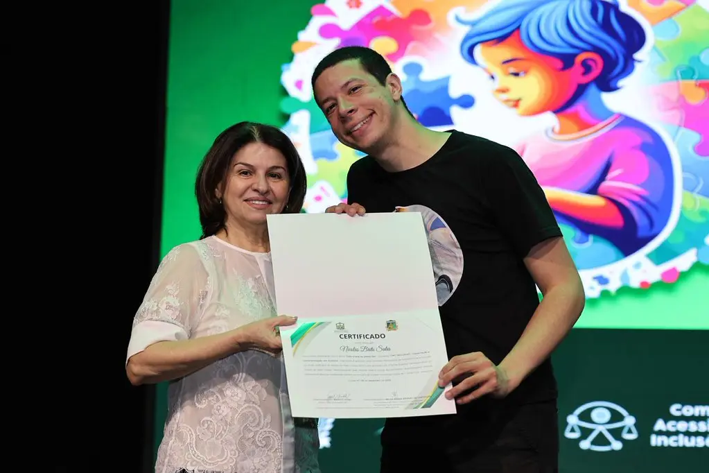 Nicolas posa ao lado da juíza Valdeci, que segura certificado. Ambos sorriem no palco, diante de painel com ilustração infantil. A cena ocorre durante cerimônia do TJMT Inclusivo em Cuiabá.