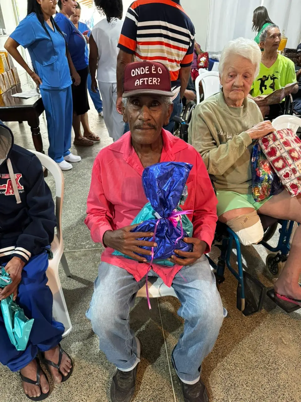 Idoso com camisa rosa e boné estampado segura um presente azul. Ao lado, uma idosa observa. Estão em cadeiras brancas, cercados de outros moradores e equipe, em ação solidária para o público idoso.