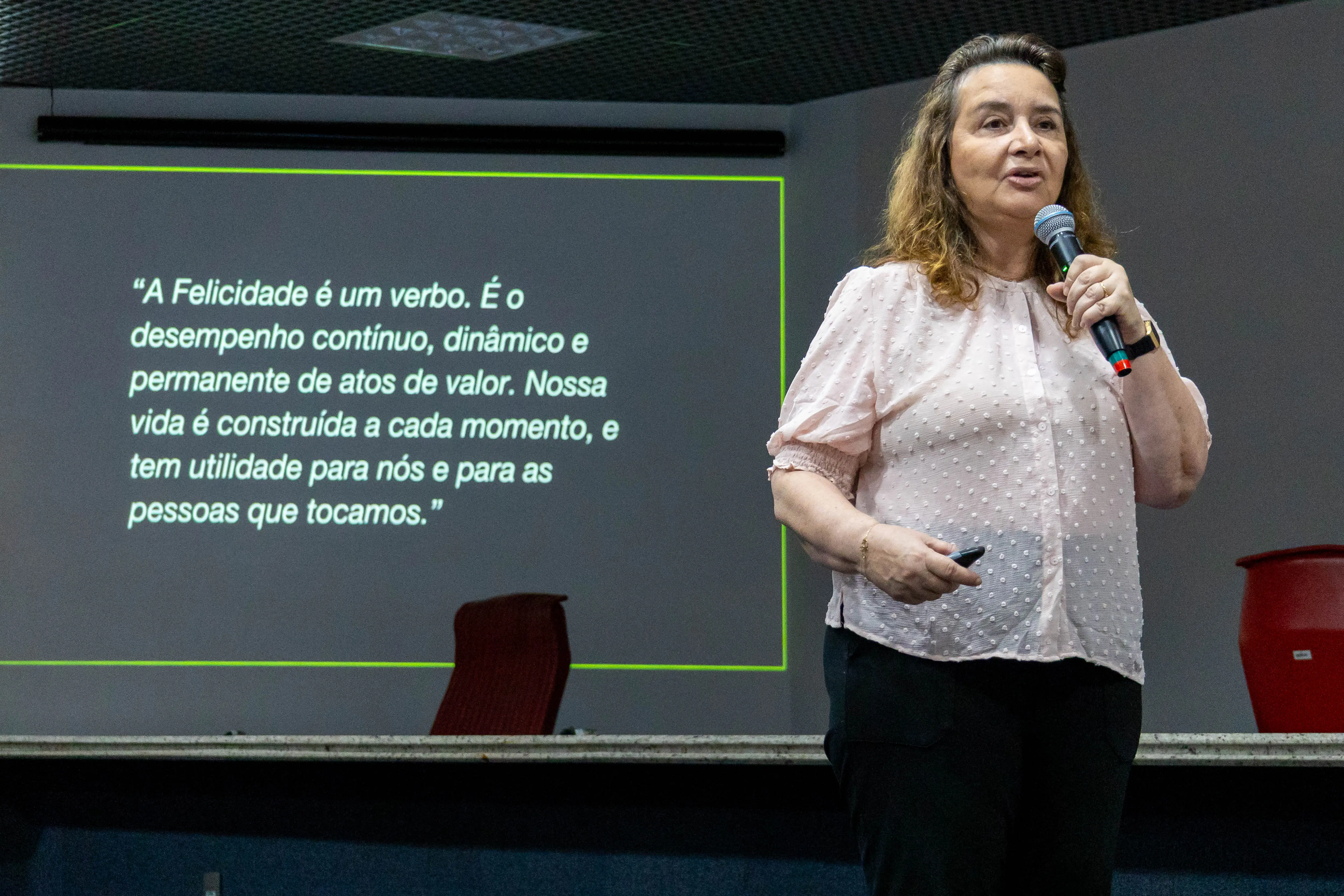 Imagem de mulher sorridente, com blusa rosa claro e calças escuras. Ela está em pé em um palco, segurando um microfone com a mão esquerda e passador de slide com a direita. No telão, atrás dela, à esquerda, é exibida em português a citação: 