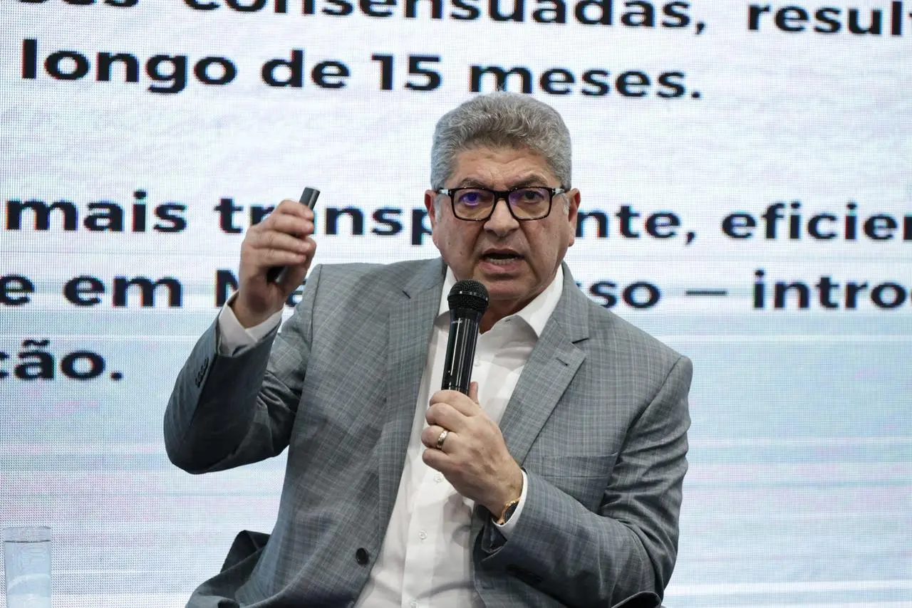 Conselheiro do Tribunal de Contas de Mato Grosso, Guilherme Maluf, sentado, falando ao microfone. Ele é um senhor branco, de cabelos grisalhos, usando camisa branca, paletó cinza e óculos de grau de armação preta. 