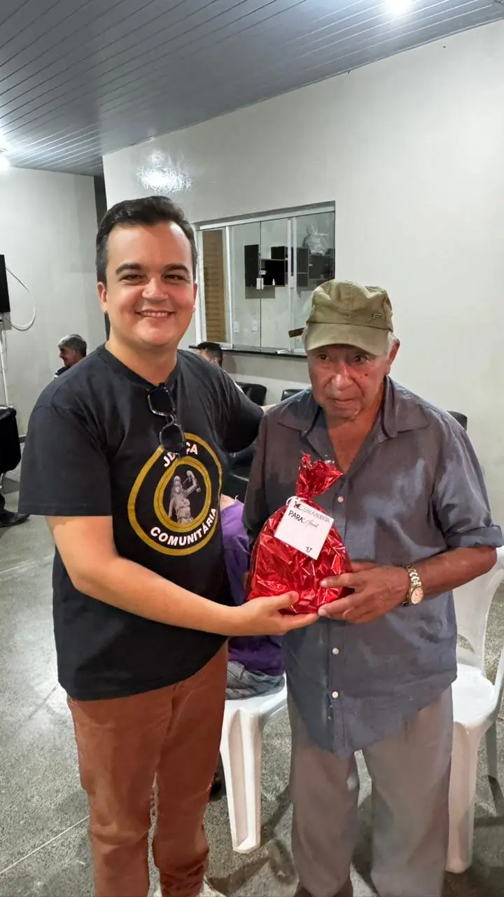 Servidor da Justiça Comunitária entrega presente vermelho a um idoso de boné bege. Eles posam sorrindo em um salão com idosos sentados ao fundo, durante distribuição de kits natalinos.