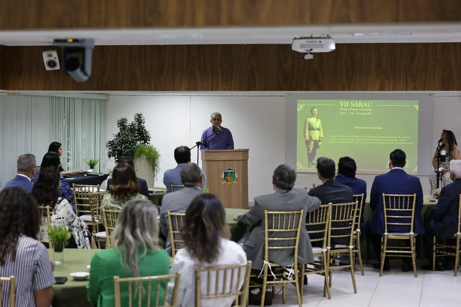 Foto de Antônio Amorim. Uma cena de evento em ambiente interno, onde um homem de camisa azul está discursando em um púlpito ao centro da sala. Uma plateia de costas, composta por várias pessoas sentadas em cadeiras de encosto dourado, assiste à apresentação.