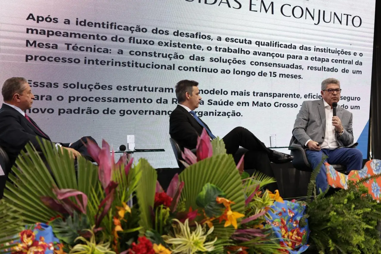 Foto que mostra os conselheiros do TCE/MT, Valter Albano e Guilherme Maluf e o juiz Agamenon Alcântara ao centro, sentados em poltronas. Maluf fala ao microfone. Atrás deles há um telão de LED e, na parte inferior da foto, há ramos de flores decorando o palco.