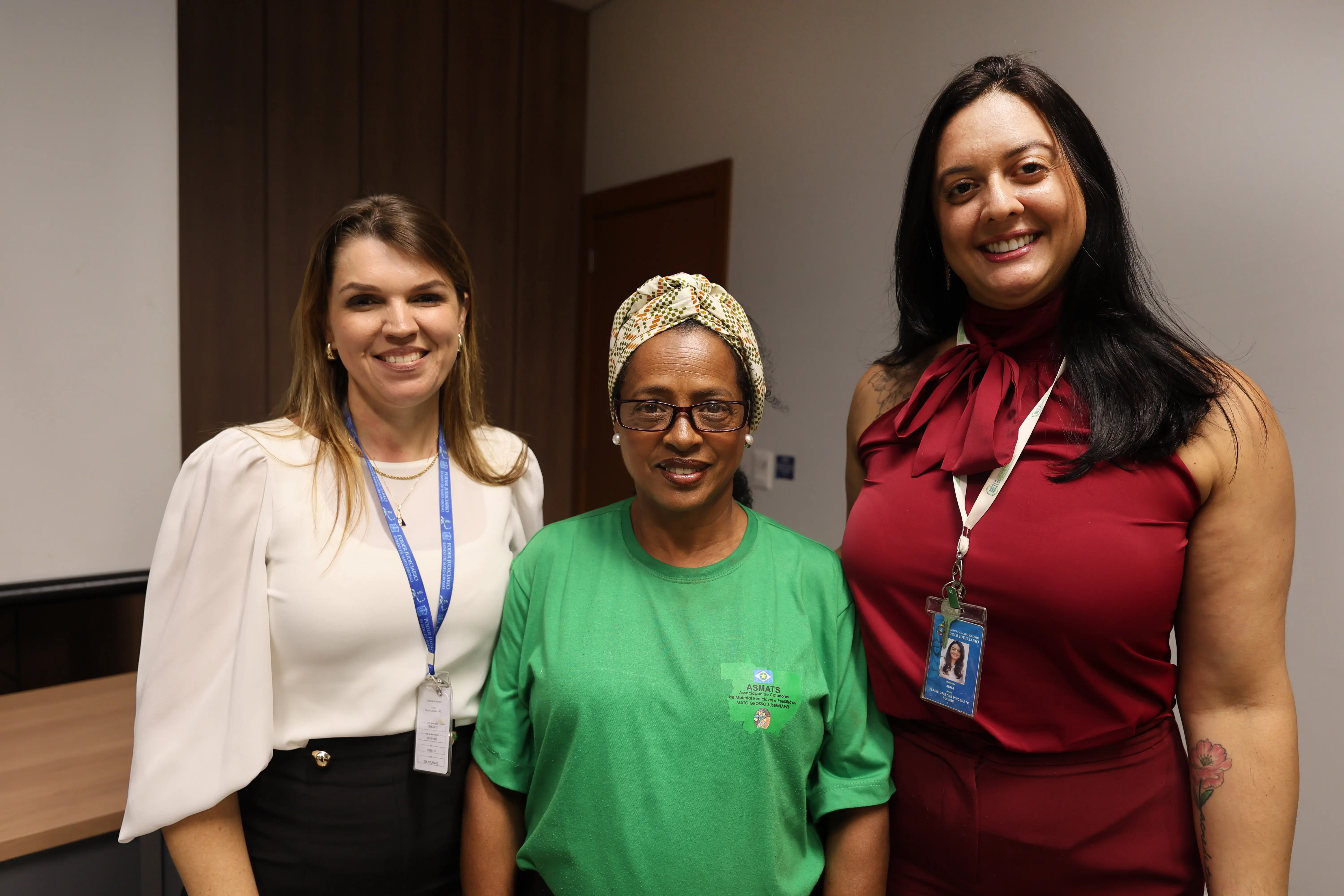 Três mulheres posam em sala do TJMT. À esquerda, servidora de blusa branca e crachá; ao centro, colaboradora com camiseta verde e turbante; à direita, servidora de roupa vinho. Todas sorriem em ambiente interno de apresentação, com painel ao fundo.