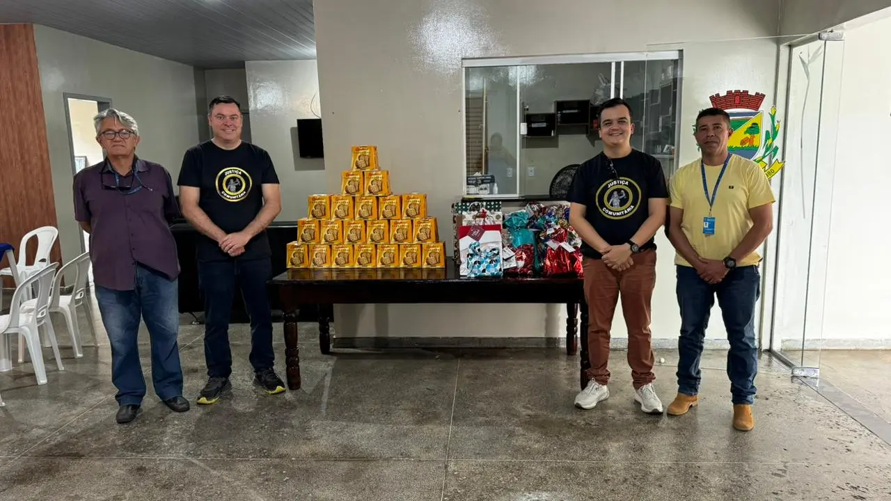 Quatro homens posam em frente a uma mesa com panetones e presentes embalados. O grupo está em ambiente interno simples, com piso de cimento e paredes claras, durante ação comunitária com idosos em Nova Brasilândia.