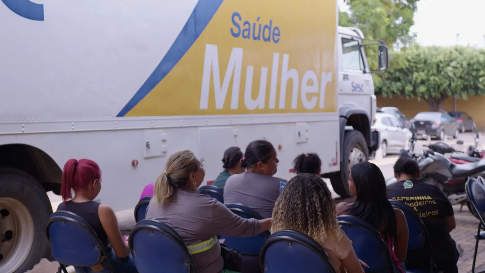 Foto que mostra várias mulheres sentadas em cadeiras ao lado da carreta Sesc Saúde Mulher.